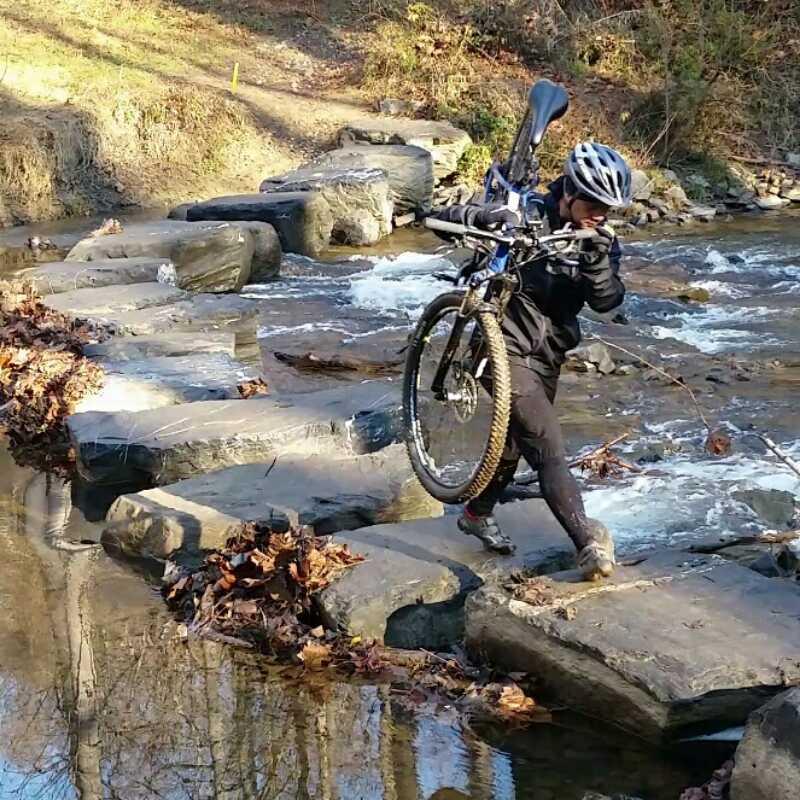 A person wearing a helmet and black clothing is carefully crossing a rocky stream while carrying a mountain bike. The water flows gently around the stones, and fallen leaves are visible on the rocks and in the water. The surrounding area features trees and greenery, indicating a natural outdoor setting. Difficult Run mountain bike trail.