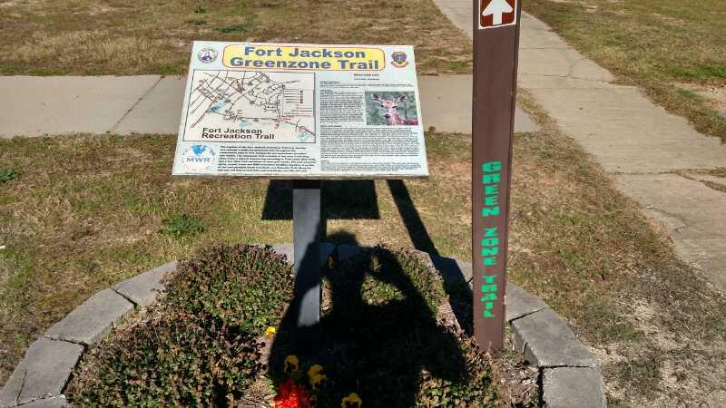 Sign for Fort Jackson Greenzone Trail next to a trailhead marker, featuring information about the recreation trail, a map, and colorful flowers at the base. The background shows a grassy area and a sidewalk. Palmetto Trail (Gate 5) mountain bike trail.