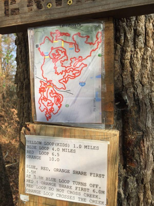 Trail map and information sign mounted on a tree. The map outlines four hiking loops: Yellow (1.0 miles), Blue (4.0 miles), Red (6.5 miles), and Orange (10.0 miles), with colored lines indicating their routes. The sign includes instructions on shared sections of the trails and notes about creek crossings for the Orange loop. Lakeshore MTB Singletrack mountain bike trail.