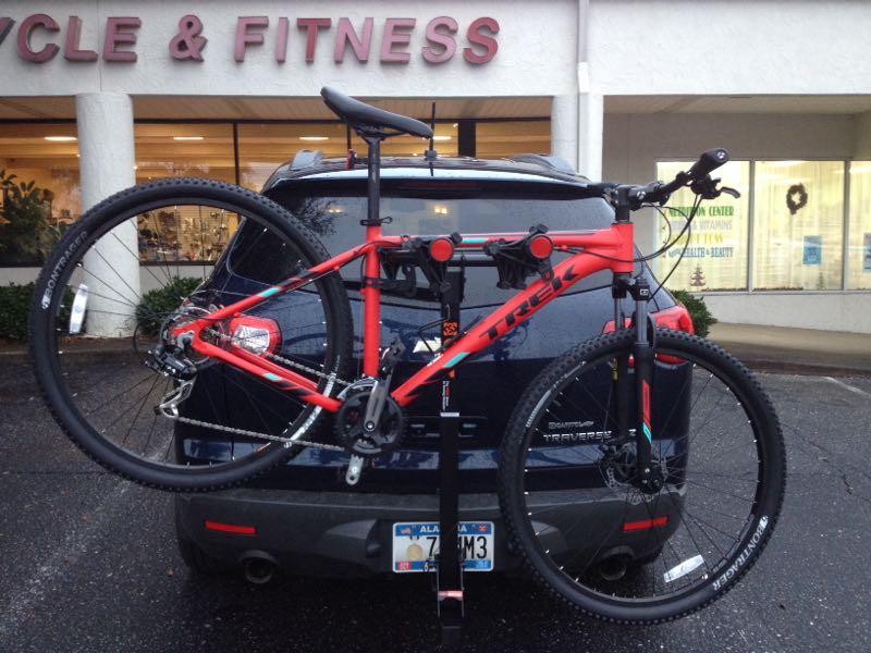 Alt text: A red Trek mountain bike is securely mounted on a bike rack attached to the back of a dark-colored SUV. The background features a store sign that reads "CYCLE & FITNESS." The setting appears to be outdoors with cloudy weather.