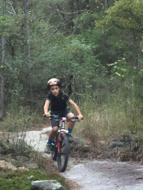 A young boy wearing a helmet rides a red bicycle on a narrow trail surrounded by greenery. He appears focused as he navigates through the forested area, with trees and shrubs in the background. Clinton Nature Preserve mountain bike trail.