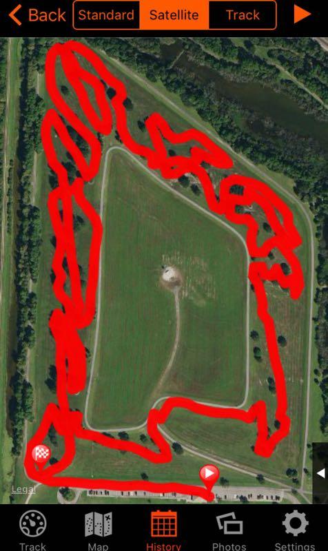 A satellite image showing a large open field with a clearly marked red path outlining the route taken, resembling a series of loops and turns. The area is surrounded by trees and a waterway, with parked cars visible near the bottom of the image. Dyer Park mountain bike trail.