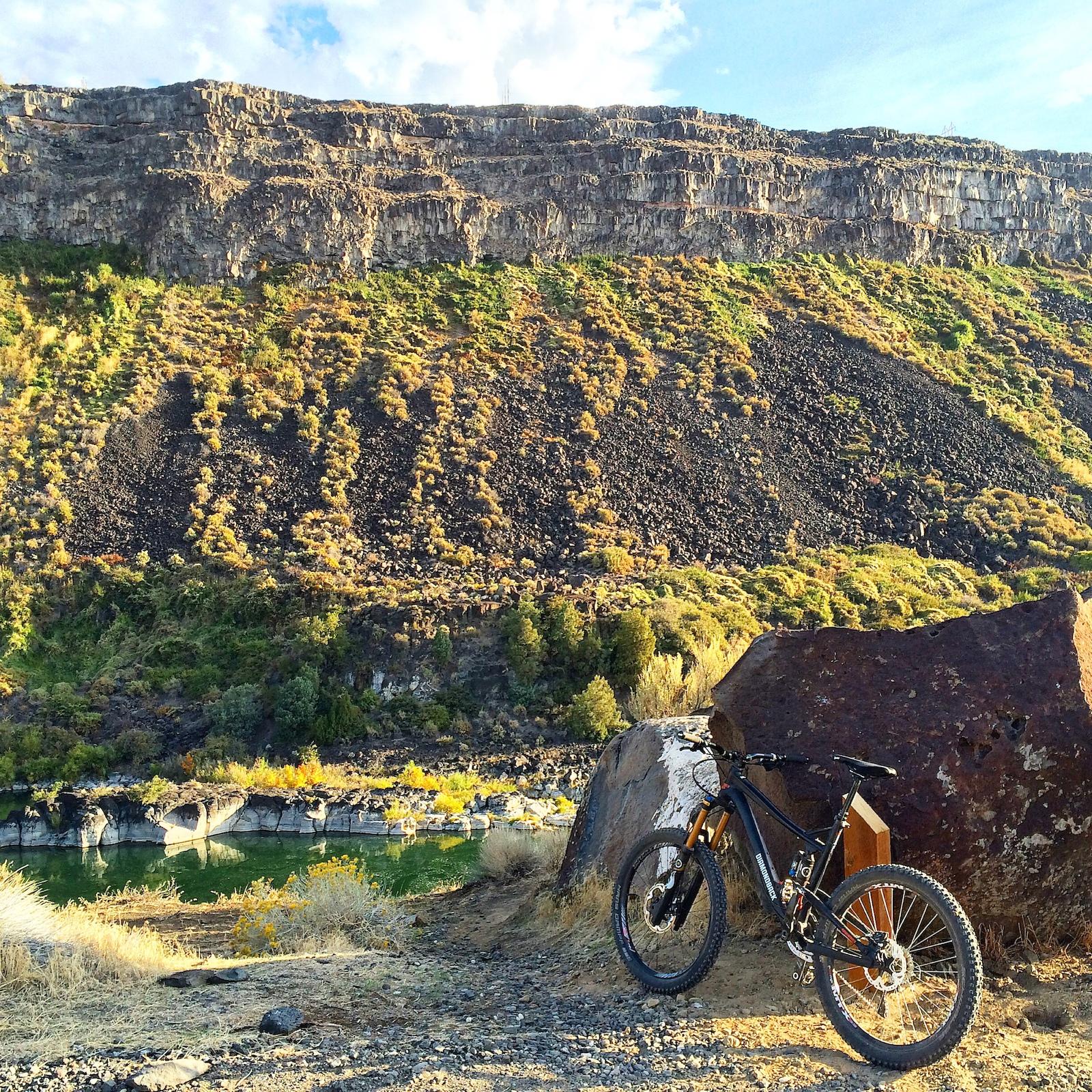 Diamondback Scapegoat: Mountain bike resting against a large rock near a riverbank, with a backdrop of steep, rocky cliffs and lush vegetation in the foreground. The scene is illuminated by warm sunlight, highlighting the diverse textures of the landscape.
