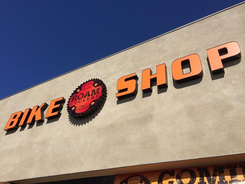 Signage for a bike shop featuring the words "BIKE SHOP" in bold orange letters against a light-colored wall, accompanied by a circular logo reading "ROAM Cyclery" with a gear design, all set against a clear blue sky.