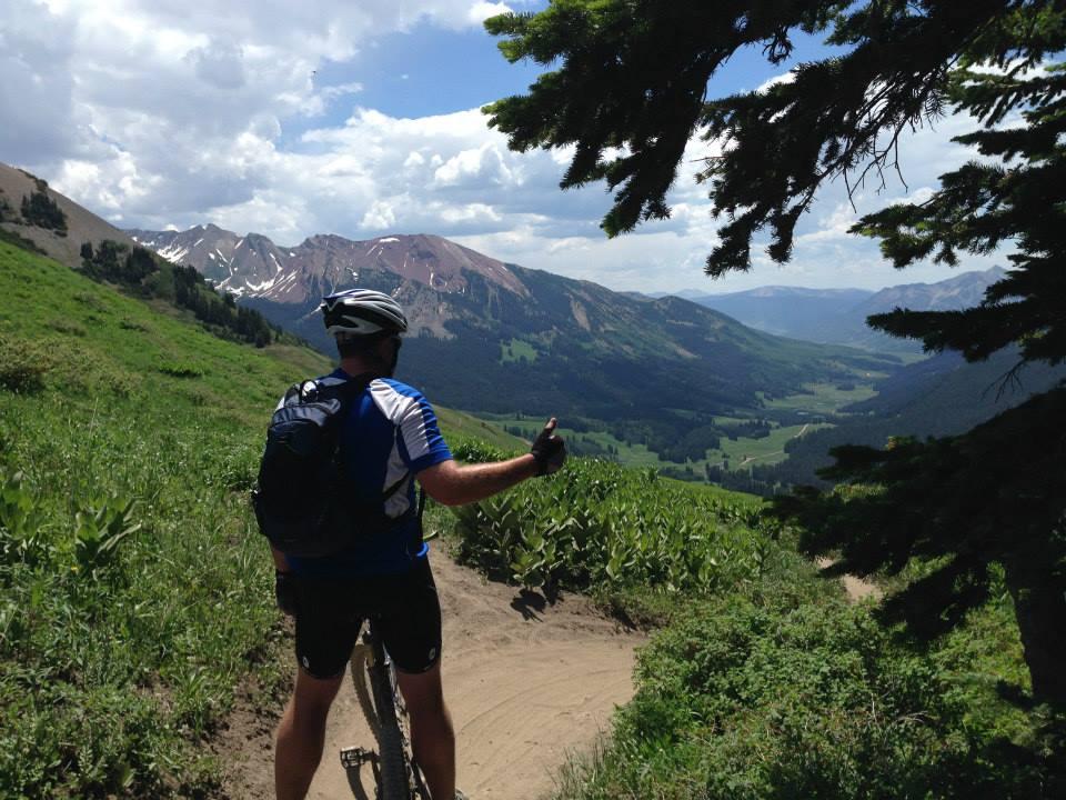 A mountain biker in a blue jersey and helmet stands on a dirt trail, giving a thumbs-up while enjoying a scenic view of green valleys and snow-capped mountains under a partly cloudy sky. Tall grass and wildflowers line the path, framed by pine trees. Trail 401 mountain bike trail.