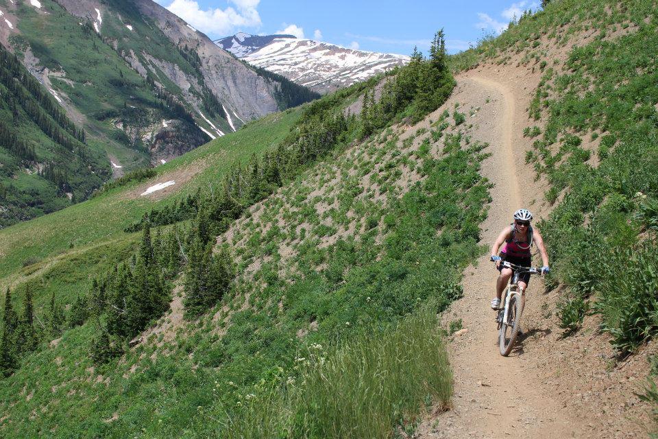 A person riding a mountain bike on a narrow dirt trail surrounded by lush green hills and distant snow-capped mountains under a clear blue sky. Trail 401 mountain bike trail.