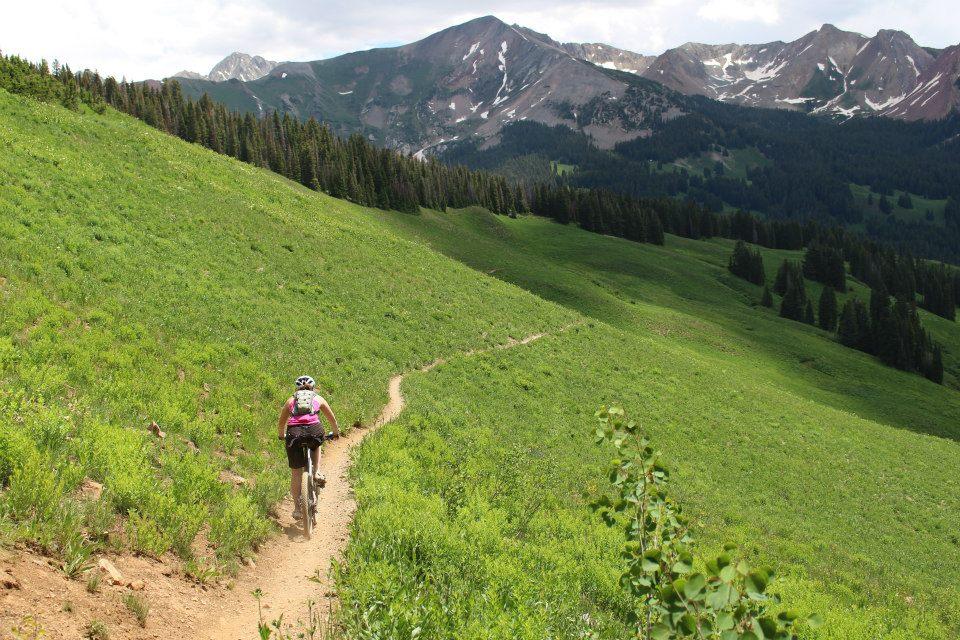 A mountain biker riding along a winding dirt trail through a lush green valley, surrounded by mountains in the background under a partly cloudy sky. Trail 401 mountain bike trail.