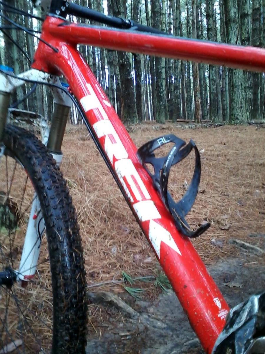 A close-up of the frame of a red Trek mountain bike, showing its muddy surface and the attached water bottle holder. The backdrop features a dense pine forest with fallen pine needles on the ground, indicating a natural outdoor setting. Carvin's Cove Trail system mountain bike trail.