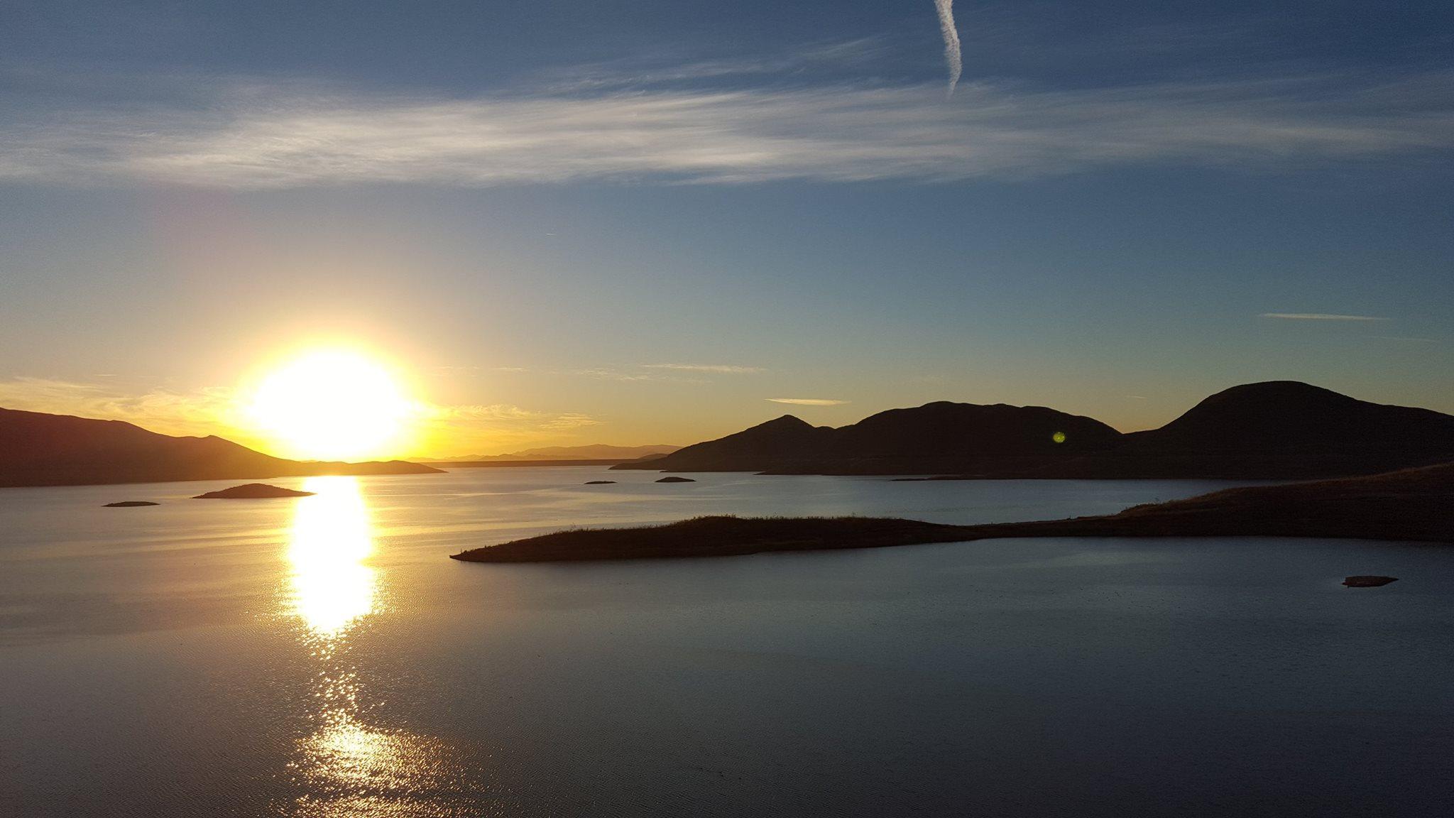 Sunset over a tranquil lake, with mountains in the background and small islands scattered across the water. The sky is painted in warm hues of orange and yellow, reflecting on the surface of the calm water. Light clouds softly float above, adding to the serene atmosphere. Lakeview Trail mountain bike trail.