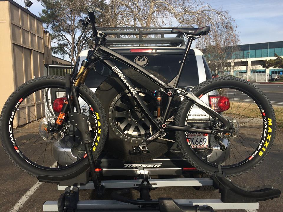 Ellsworth Epiphany 275 Enduro: A black mountain bike mounted on a bike rack attached to the back of a vehicle. The bike features large tires and a sleek frame, with visible branding on the wheels. The background includes a parking lot and a building, suggesting an outdoor setting.