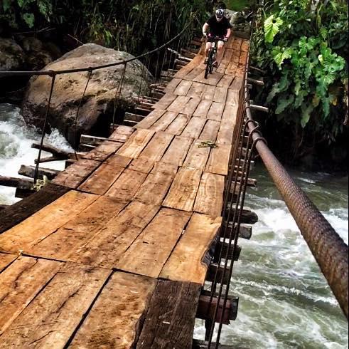 A person riding a bicycle on a narrow wooden bridge suspended over a flowing river, surrounded by lush greenery and rocks. Via a Cunuco mountain bike trail.