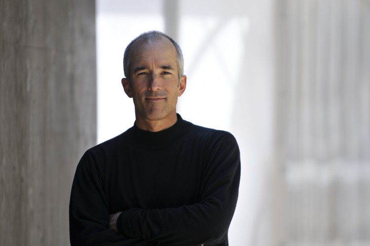 A man with short gray hair wearing a black turtleneck sweater stands confidently with his arms crossed. The background features soft, blurred architectural elements, suggesting an indoor or urban setting with natural light.