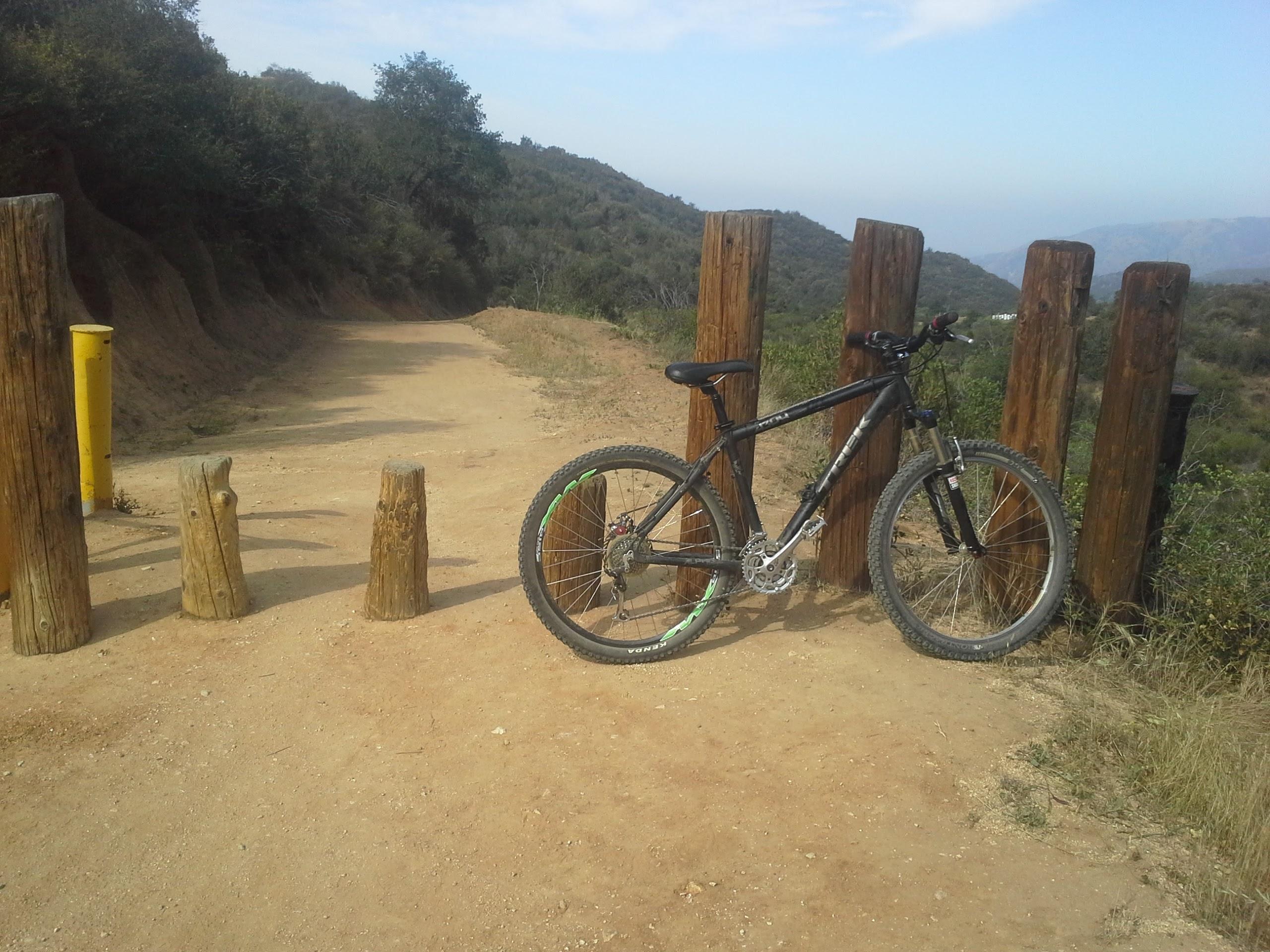 A black mountain bike resting against wooden posts on a dirt trail, with a scenic view of hills and trees in the background under a clear sky. Marshall Canyon Park mountain bike trail.