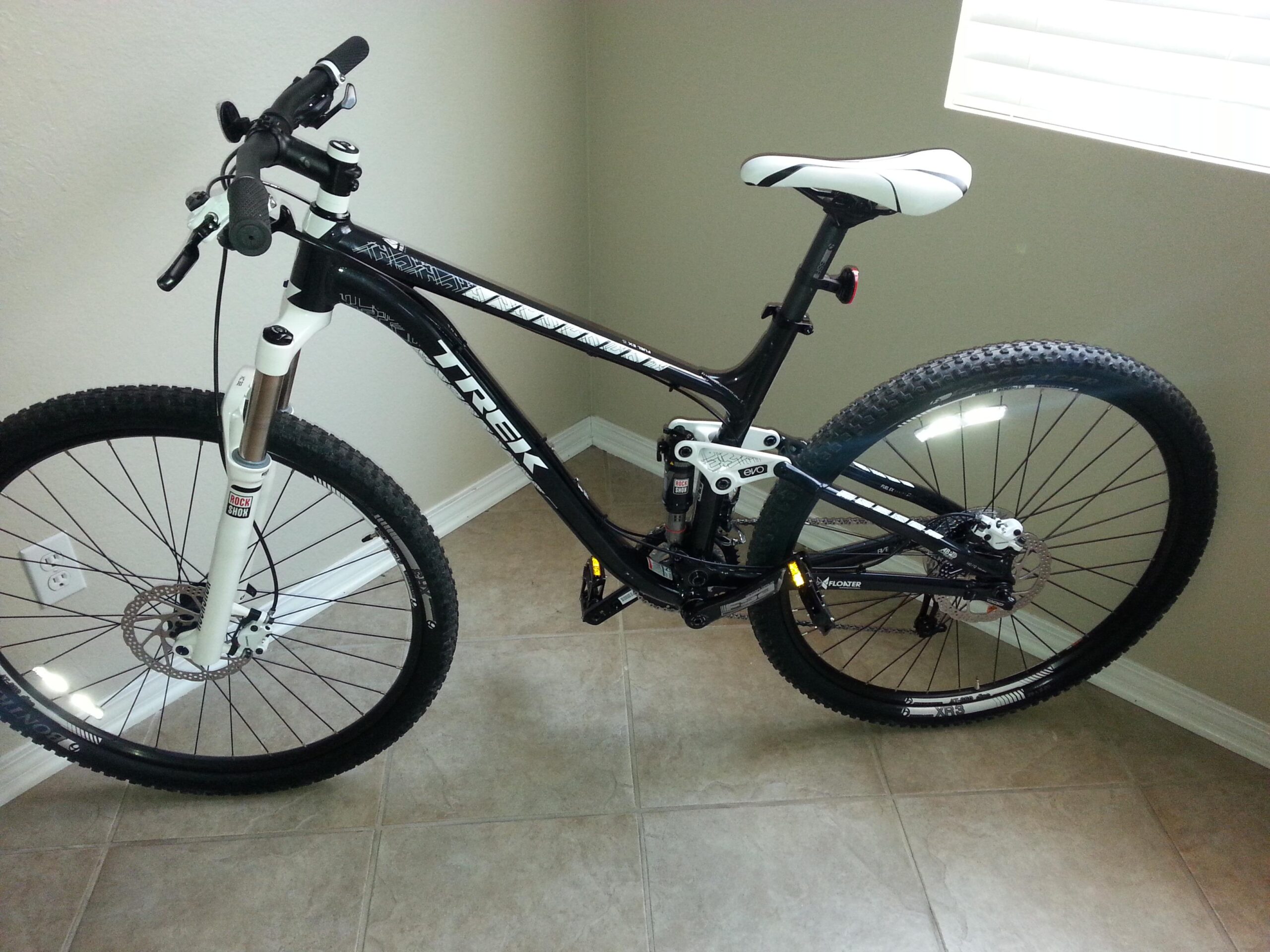 Trek Fuel EX 5 29: Mountain bike leaning against a wall, featuring a black frame with white accents, large tires, and suspension systems. The bike is positioned on tiled flooring in a well-lit room with a window in the background.