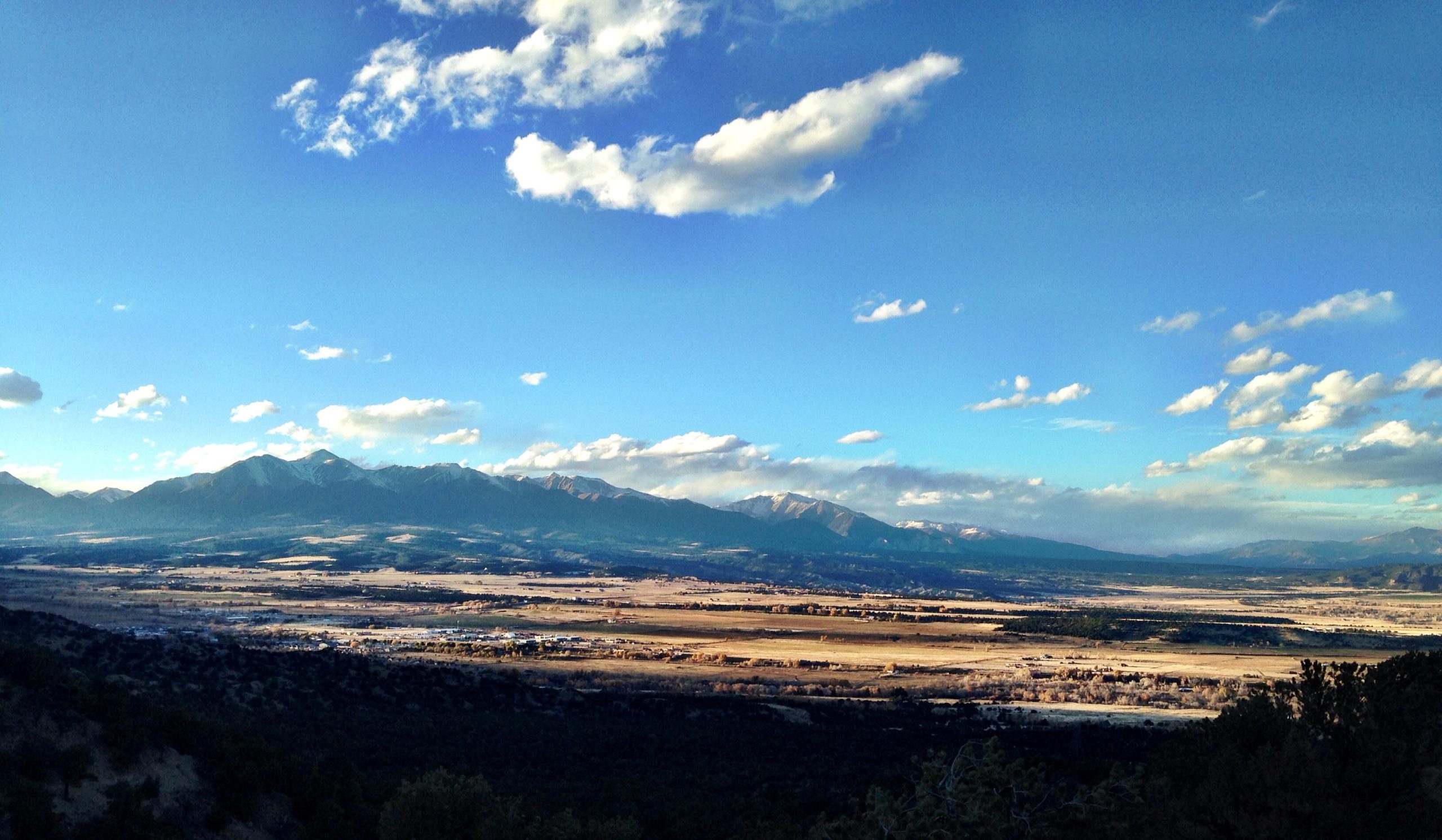 A panoramic view of a mountainous landscape under a bright blue sky with scattered clouds. The foreground features a valley with patches of land, while snow-capped peaks rise majestically in the background, creating a serene and picturesque natural scene. Double Rainbow mountain bike trail.