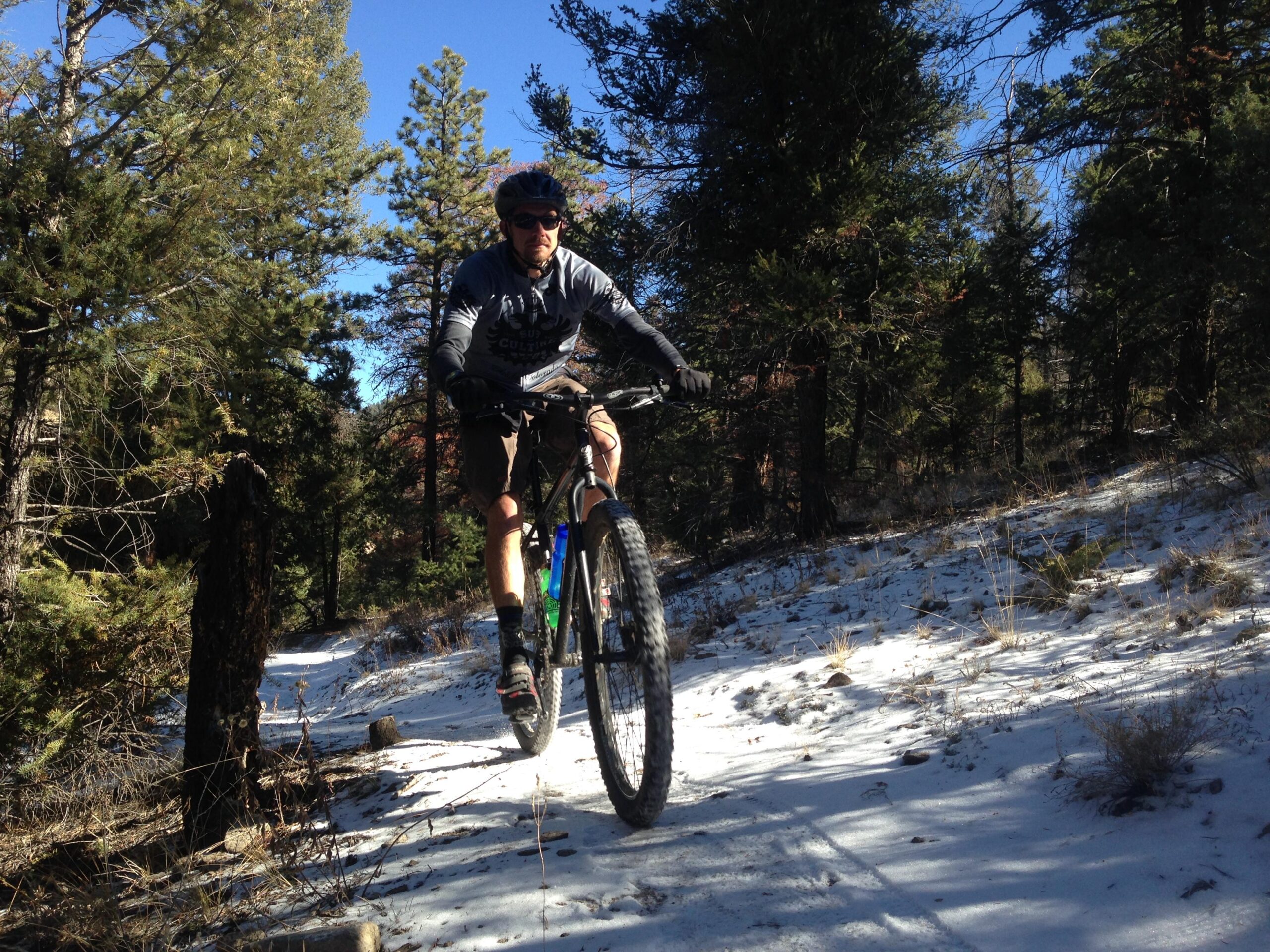 A mountain biker riding on a snowy trail surrounded by pine trees, wearing a helmet and sunglasses. The rider is mid-action, with the bike's front wheel lifted off the ground. The scene is set in a forest with a clear blue sky above. Cottonwood mountain bike trail.