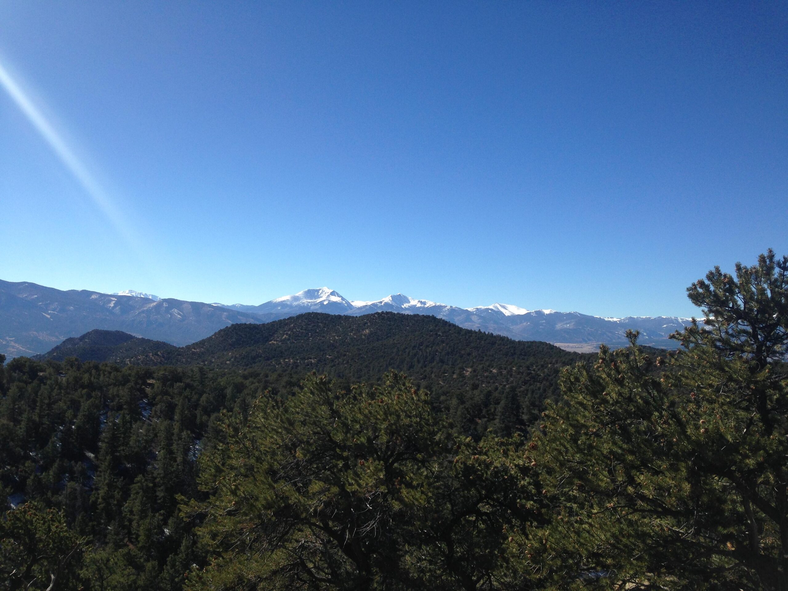 A scenic view of mountainous terrain under a clear blue sky, featuring snow-capped peaks in the distance. The foreground includes lush green trees and rolling hills, creating a tranquil natural landscape. Cottonwood mountain bike trail.