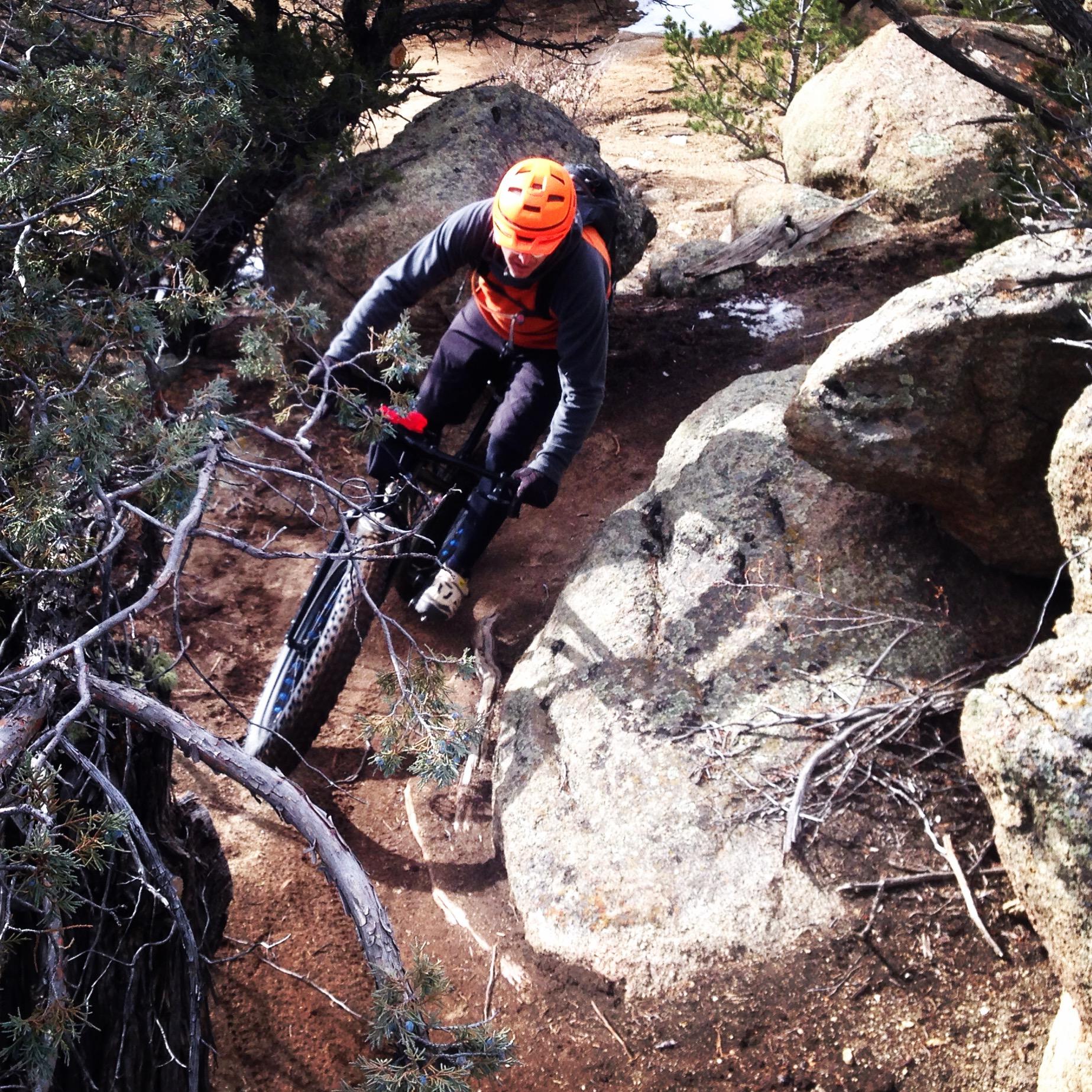 A mountain biker navigates a rocky trail, leaning into a turn. The rider wears a bright orange helmet and an orange vest, surrounded by trees and large boulders. The terrain is dusty with some scattered branches. Midland Hills Trails mountain bike trail.