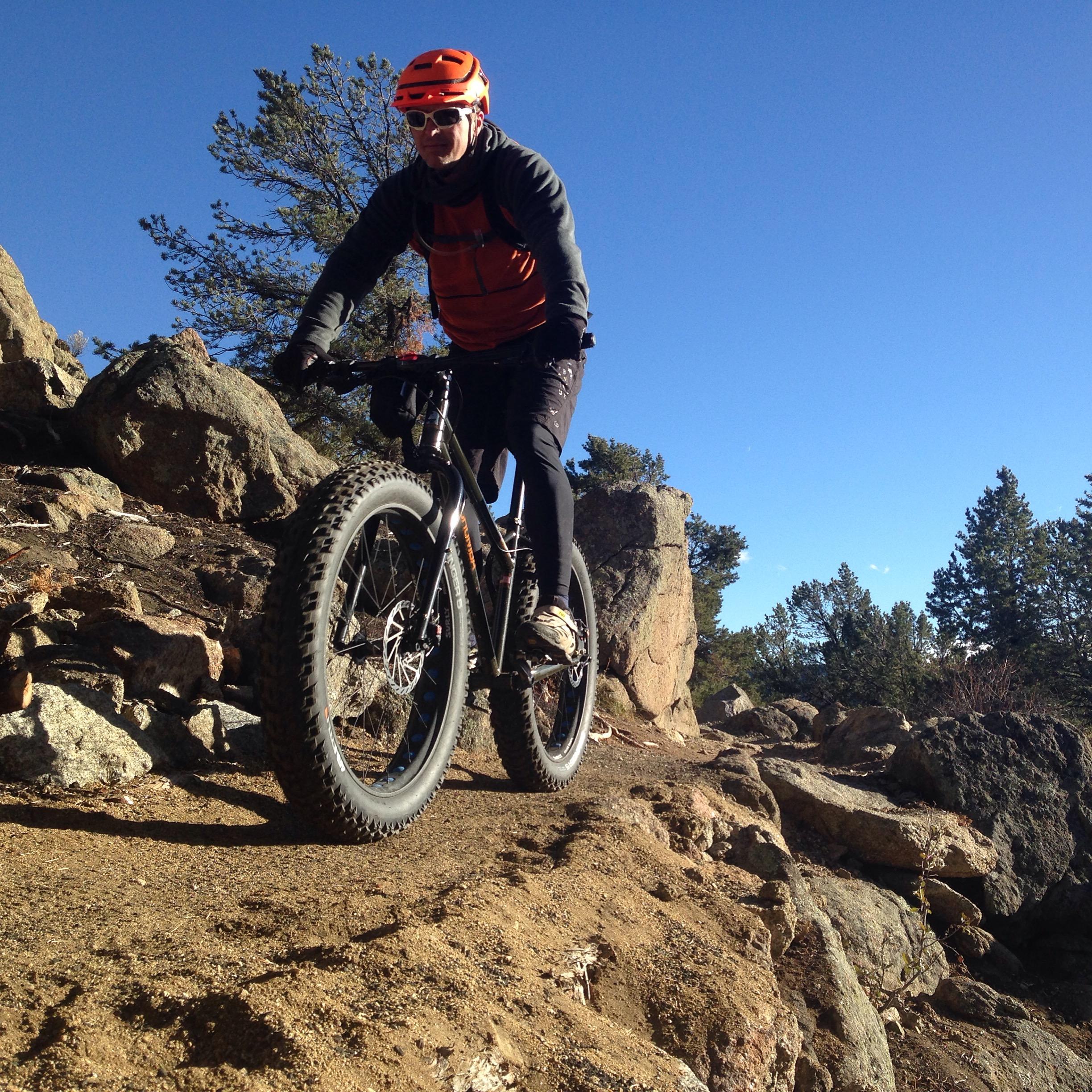 A mountain biker riding on a rocky trail, wearing an orange helmet and black clothing, with a clear blue sky and trees in the background. Midland Hills Trails mountain bike trail.