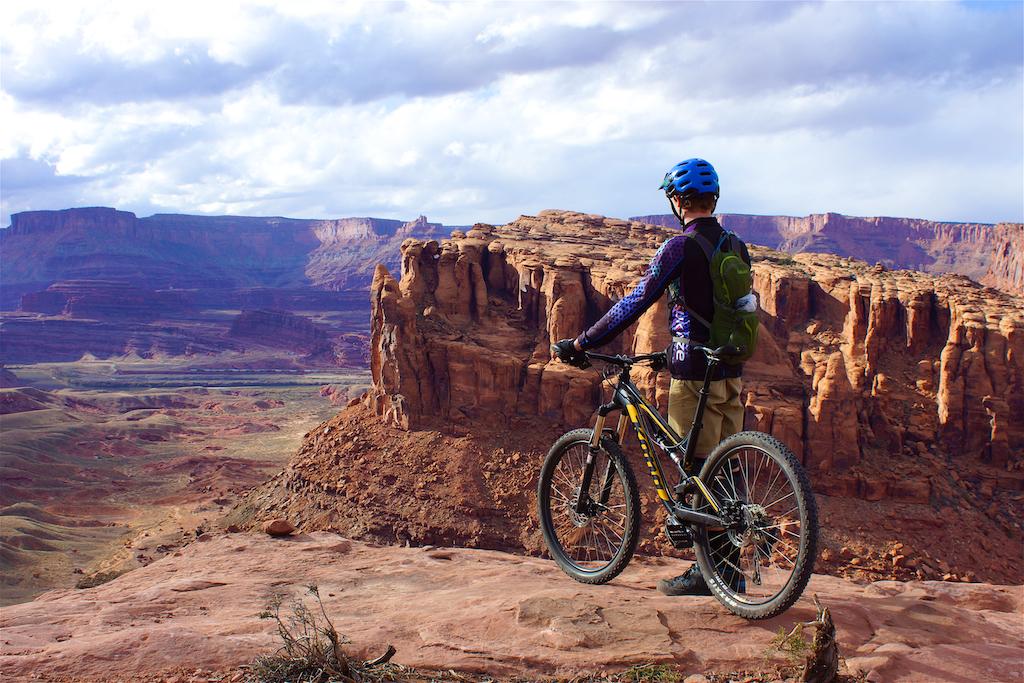 A mountain biker standing on a rocky ledge, overlooking a vast canyon landscape with red rock formations and rolling hills under a partly cloudy sky. The biker is wearing a blue helmet, a long-sleeve shirt, and has a backpack. Captain Ahab mountain bike trail.