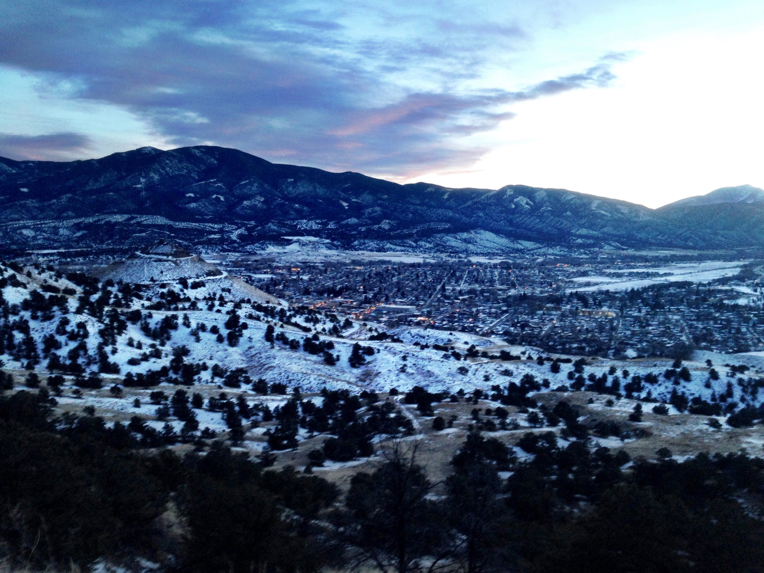 A scenic view of a snow-covered landscape featuring rolling hills and mountains under a twilight sky. The town below is illuminated with warm lights, contrasting with the cool tones of the snow and the dark silhouettes of the trees scattered throughout the foreground. North Backbone mountain bike trail.