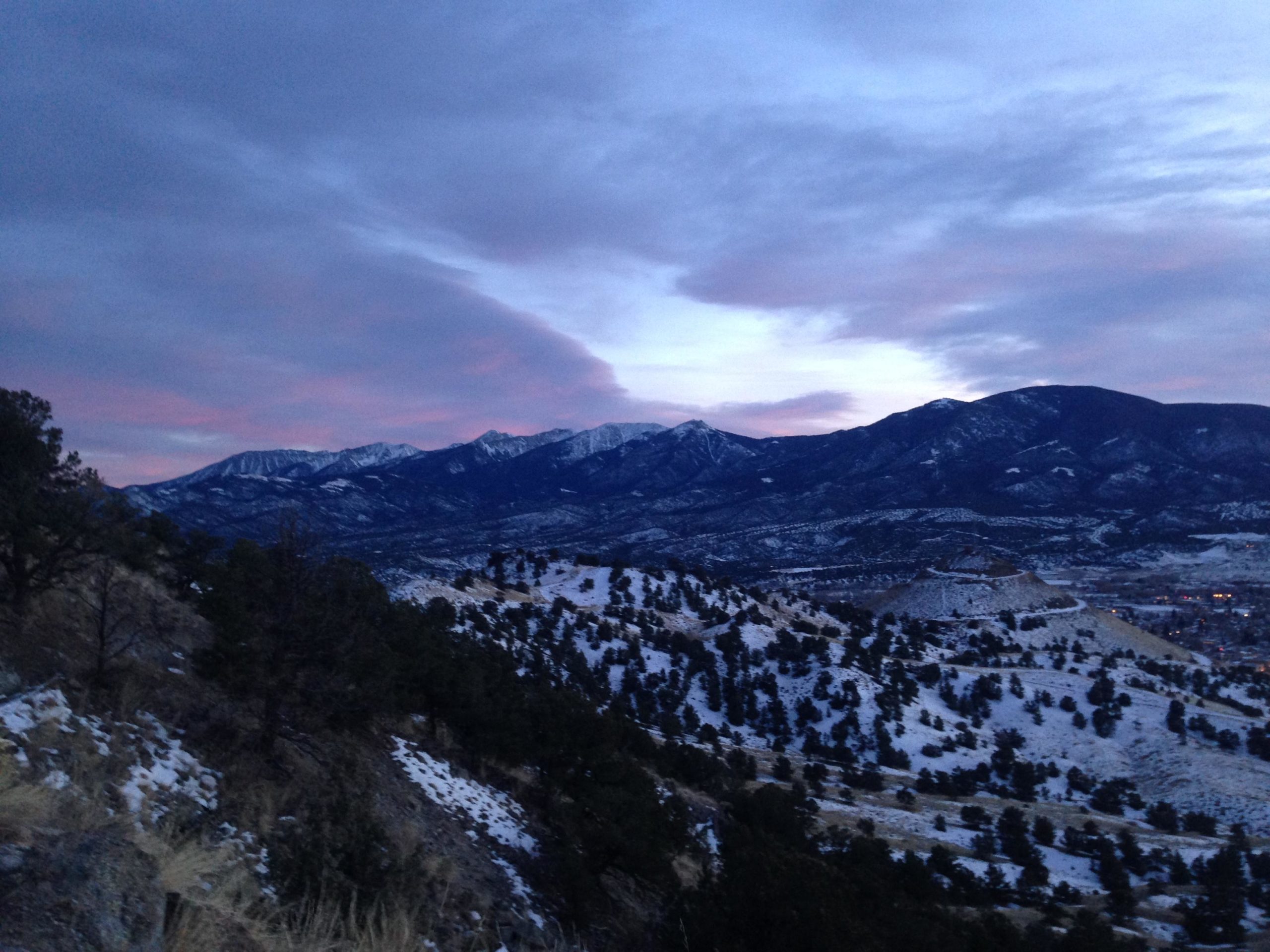 A scenic view of snow-covered mountains during twilight, featuring a gradient sky with shades of purple and blue. The foreground includes rocky terrain and sparse pine trees, while the mountains in the background are partially obscured by clouds. North Backbone mountain bike trail.