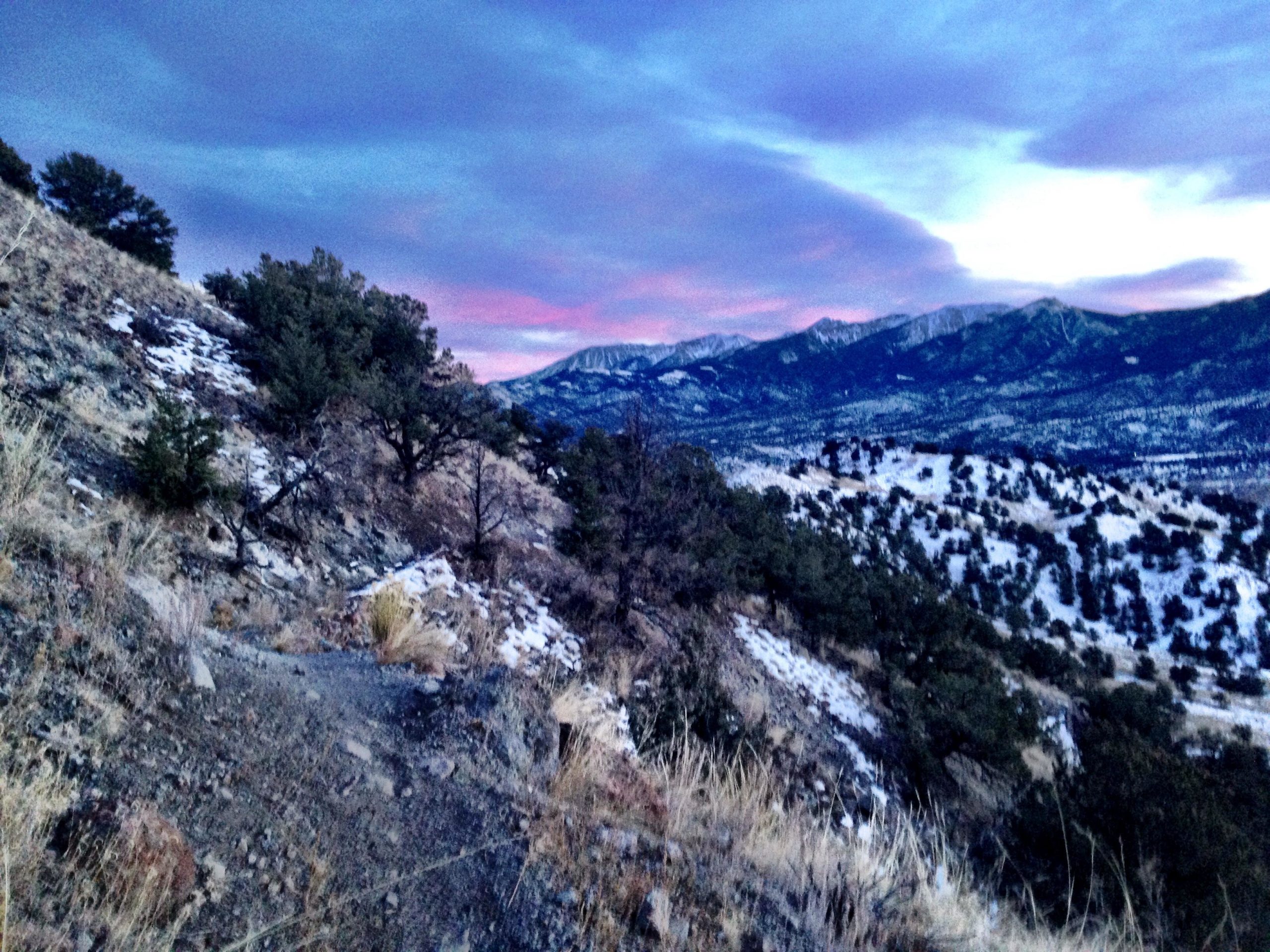 A winding dirt path along a rugged mountainside, surrounded by patches of snow and sparse vegetation. The scene is illuminated by a soft twilight glow, with colorful clouds reflecting the fading light of the sunset over distant mountain peaks. North Backbone mountain bike trail.