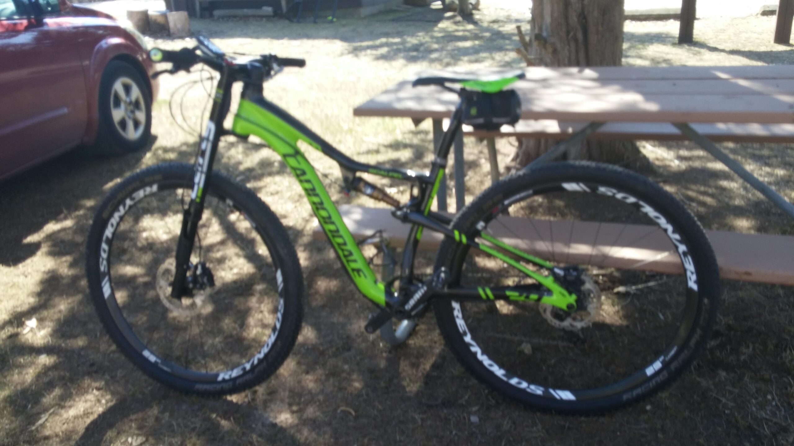 Cannondale Scalpel: A green and black mountain bike parked next to a picnic table in a grassy outdoor area, with a red car visible in the background. The bike features thick tires and disc brakes, and a small bag is attached to the seat.