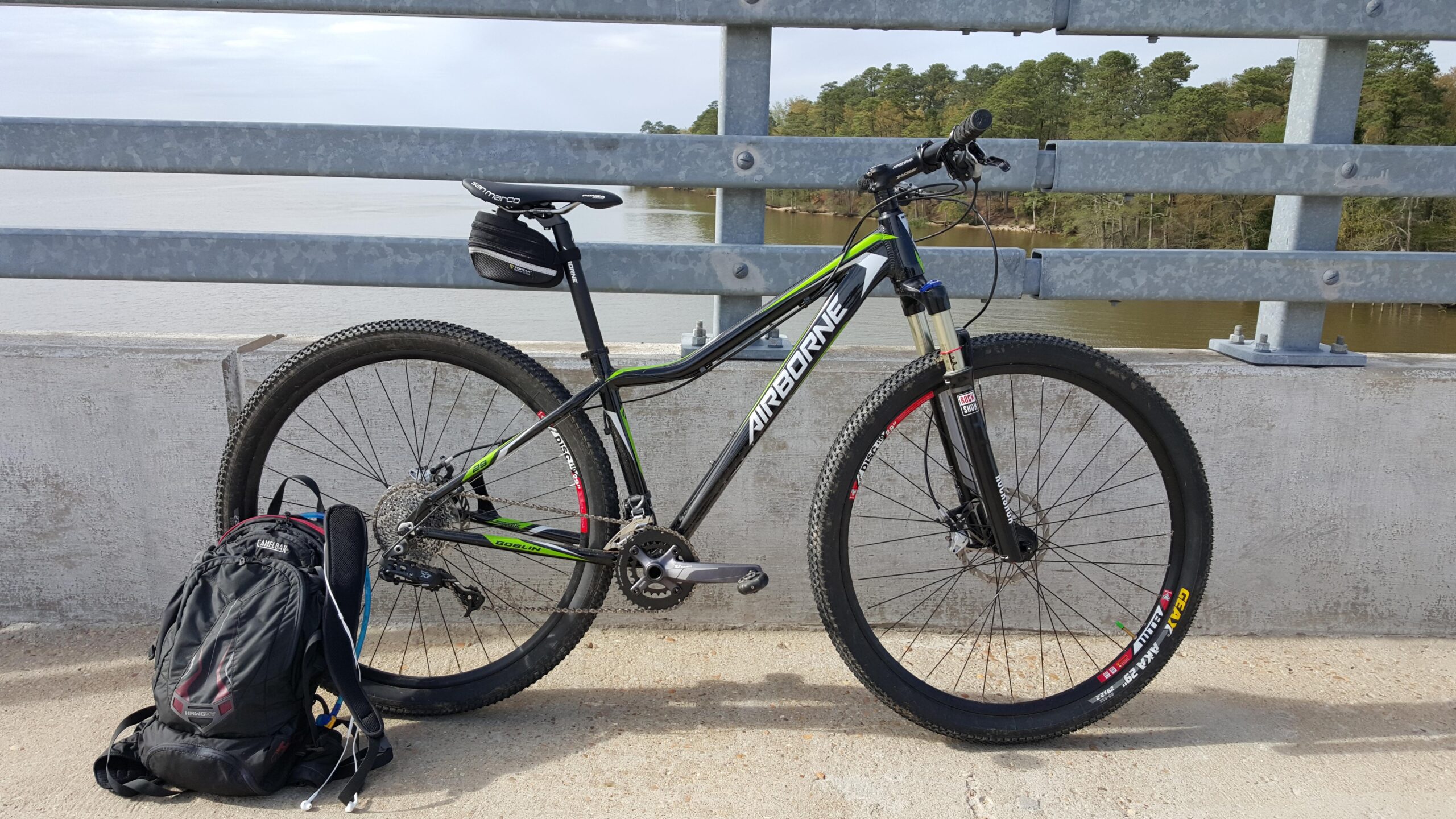 Airborne Goblin: A mountain bike with black and green accents is leaning against a railing on a bridge. A backpack is placed on the ground beside the bike, with a view of a lake and trees in the background. The scene is captured on a sunny day.