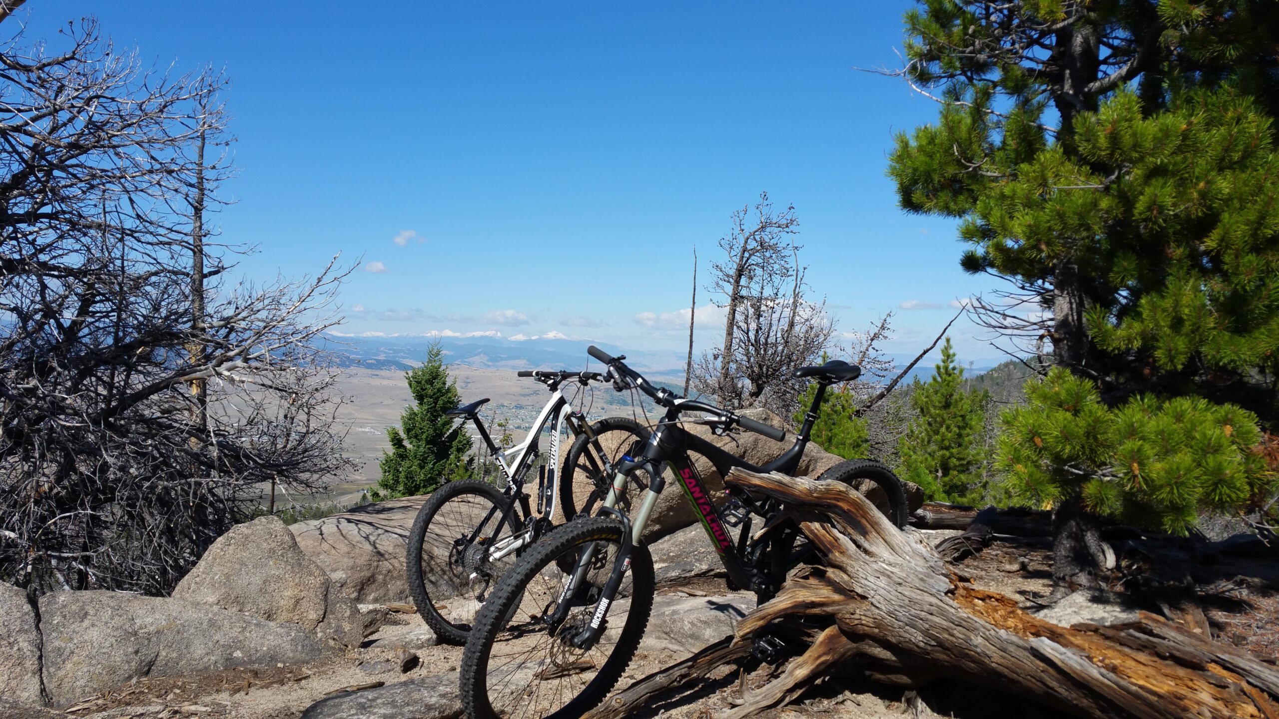 Santa Cruz Bronson: Two mountain bikes are parked on rocky terrain, framed by a mix of green and dead trees. In the background, a panoramic view showcases rolling hills and distant mountains under a clear blue sky. The scene captures a sense of adventure and nature.