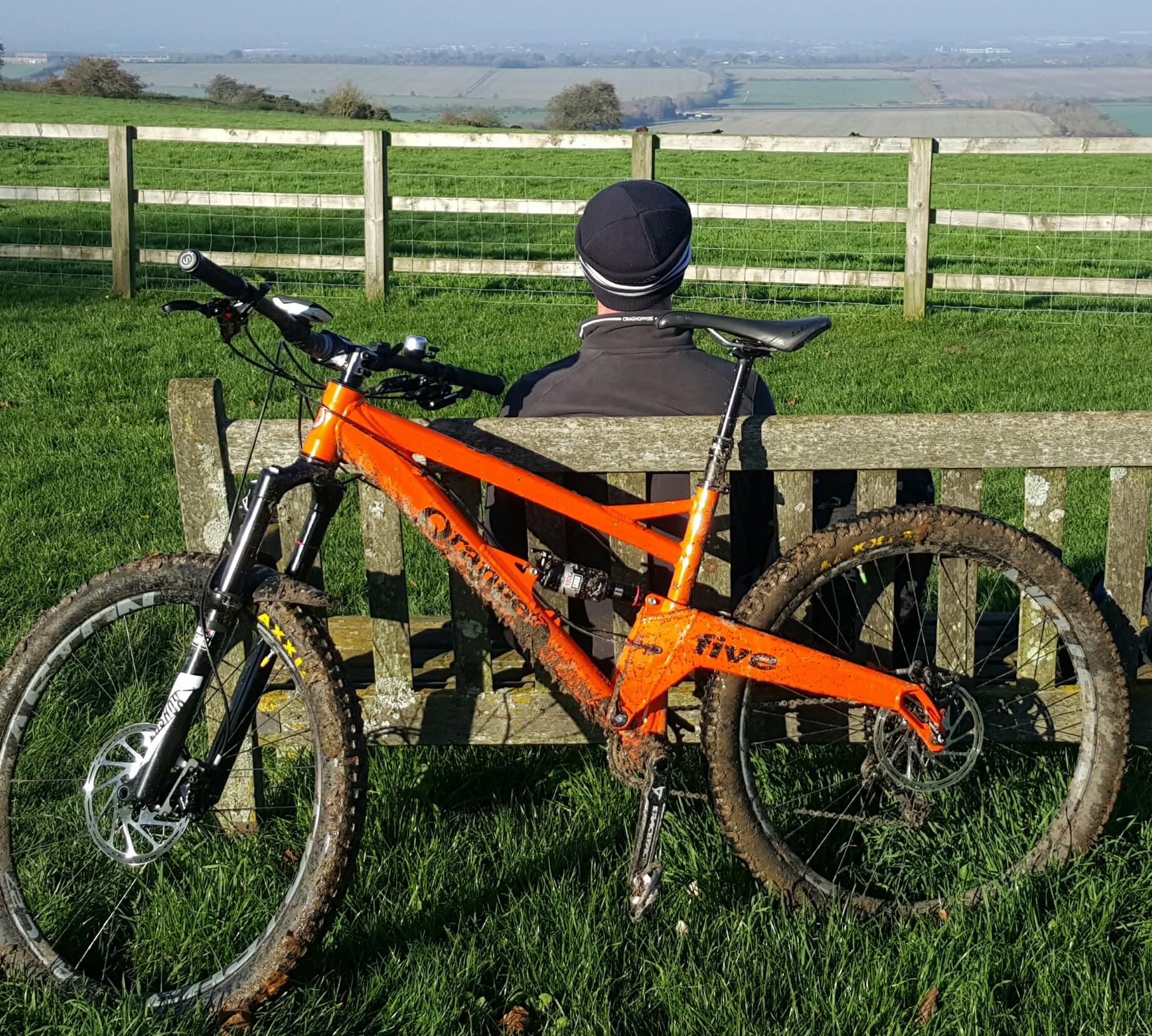 Orange Five RS: A person sitting on a wooden bench with their back turned, overlooking a scenic view of green fields and distant hills. An orange mountain bike, partially covered in mud, leans against the bench next to them. The scene is bathed in sunlight, suggesting a clear, pleasant day.