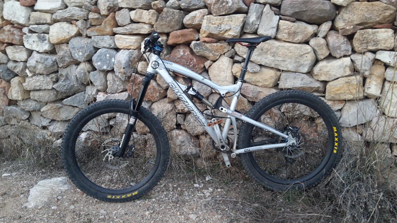 Santa Cruz Nomad: A silver mountain bike with thick tires is leaning against a rustic stone wall, surrounded by sparse grass and gravel. The bike features prominent branding and a suspension system, suited for off-road riding.