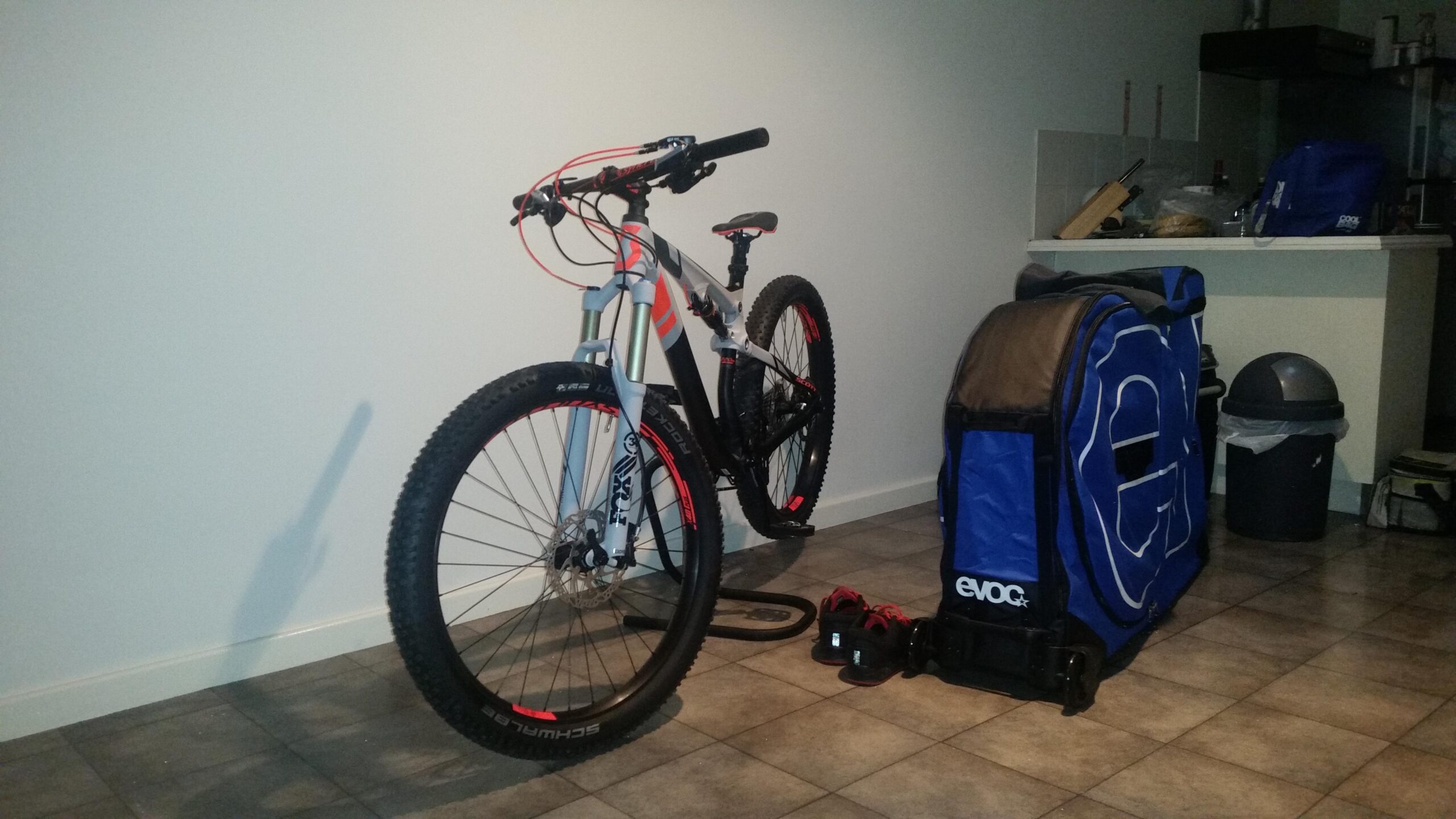 Scott Genius Plus: A mountain bike with red and black accents is leaning against a wall in a room with tiled flooring. Next to the bike, there is a large blue travel bag, a pair of shoes, and a trash can in the background. The setting appears to be indoors, with kitchen items visible on a counter in the distance.