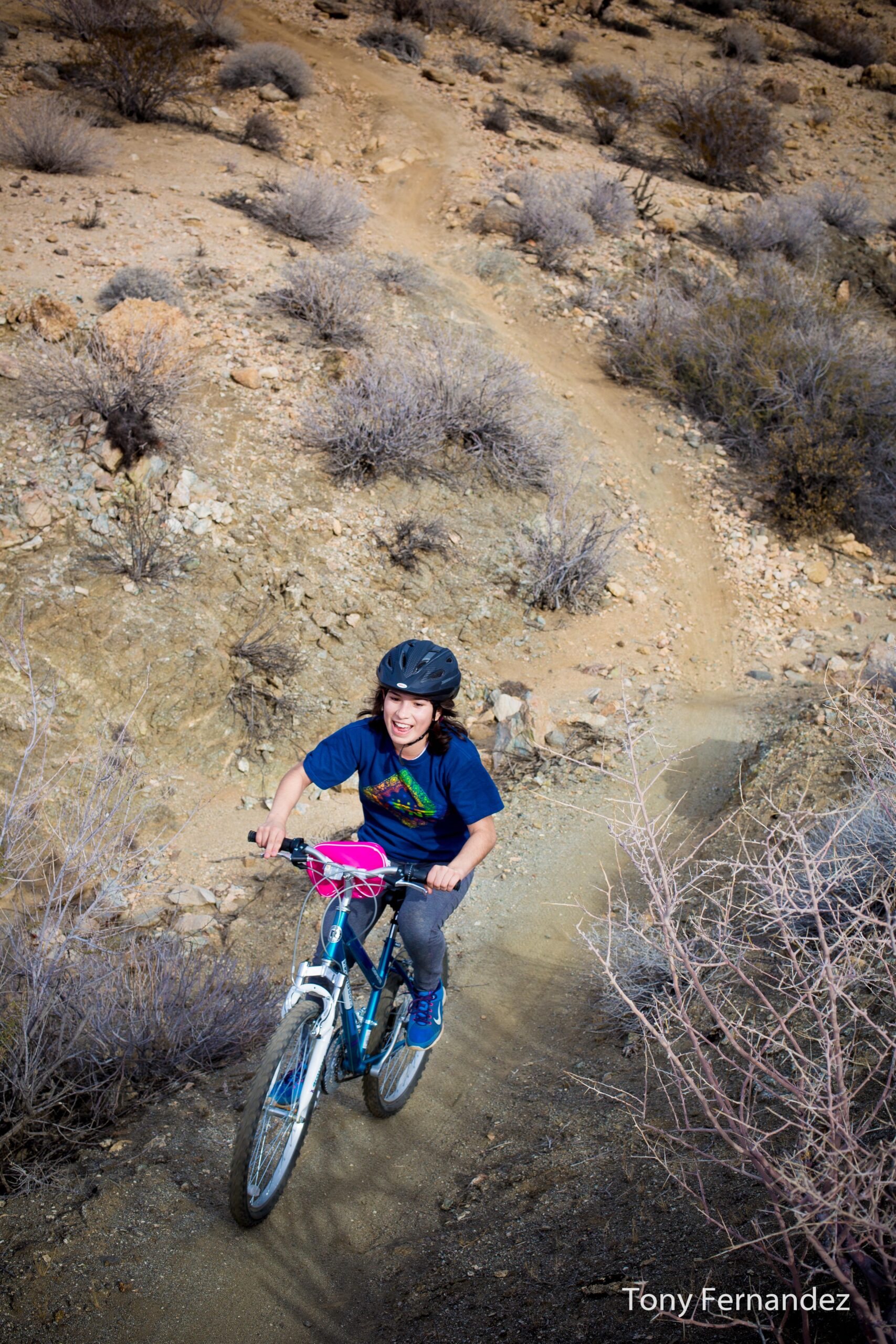 A young girl wearing a helmet rides a blue mountain bike on a dirt trail, smiling as she navigates a hilly, rocky landscape dotted with dry bushes. Dunn Road: Hahn / Buena Vista mountain bike trail.