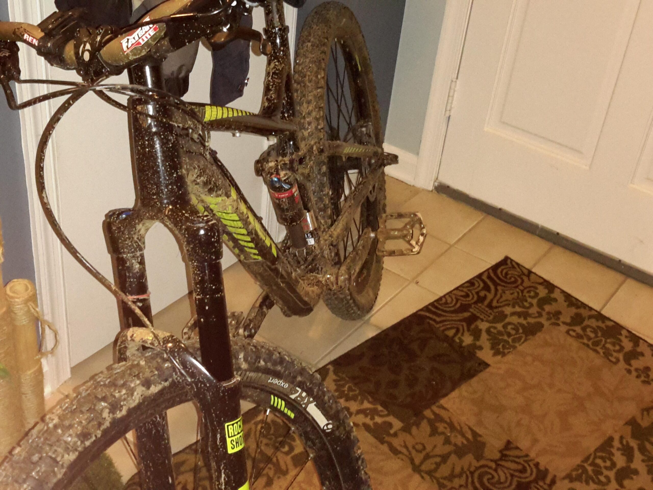 Trek Trek Remedy 8 27.5: Mountain bike covered in mud parked inside a home, next to a decorative rug and a closed door. The bike features a black frame with green accents and visible dirt on the tires and components.