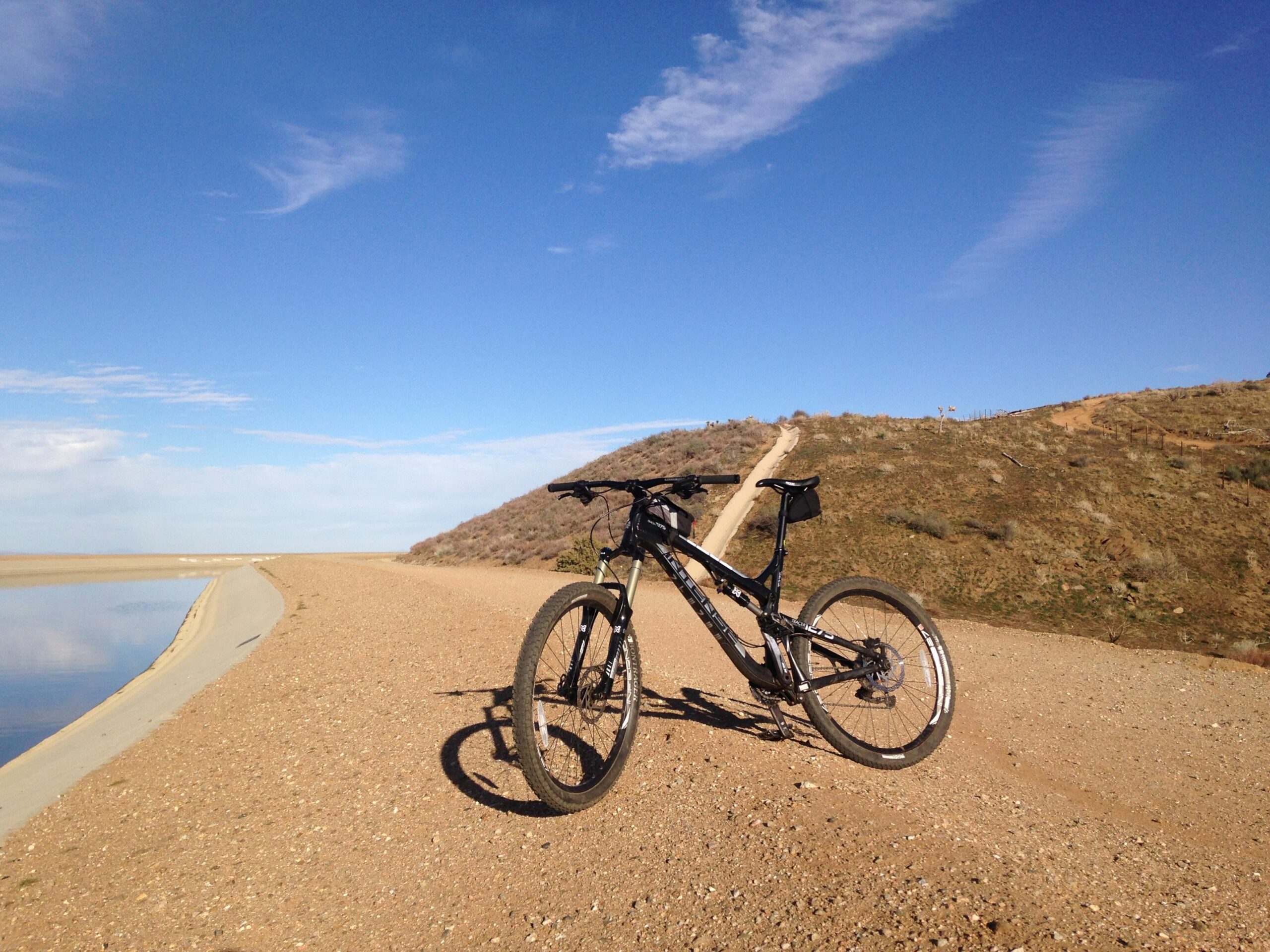 Intense tracer: A black mountain bike resting on a gravel path beside a calm body of water, with a hill in the background under a clear blue sky.