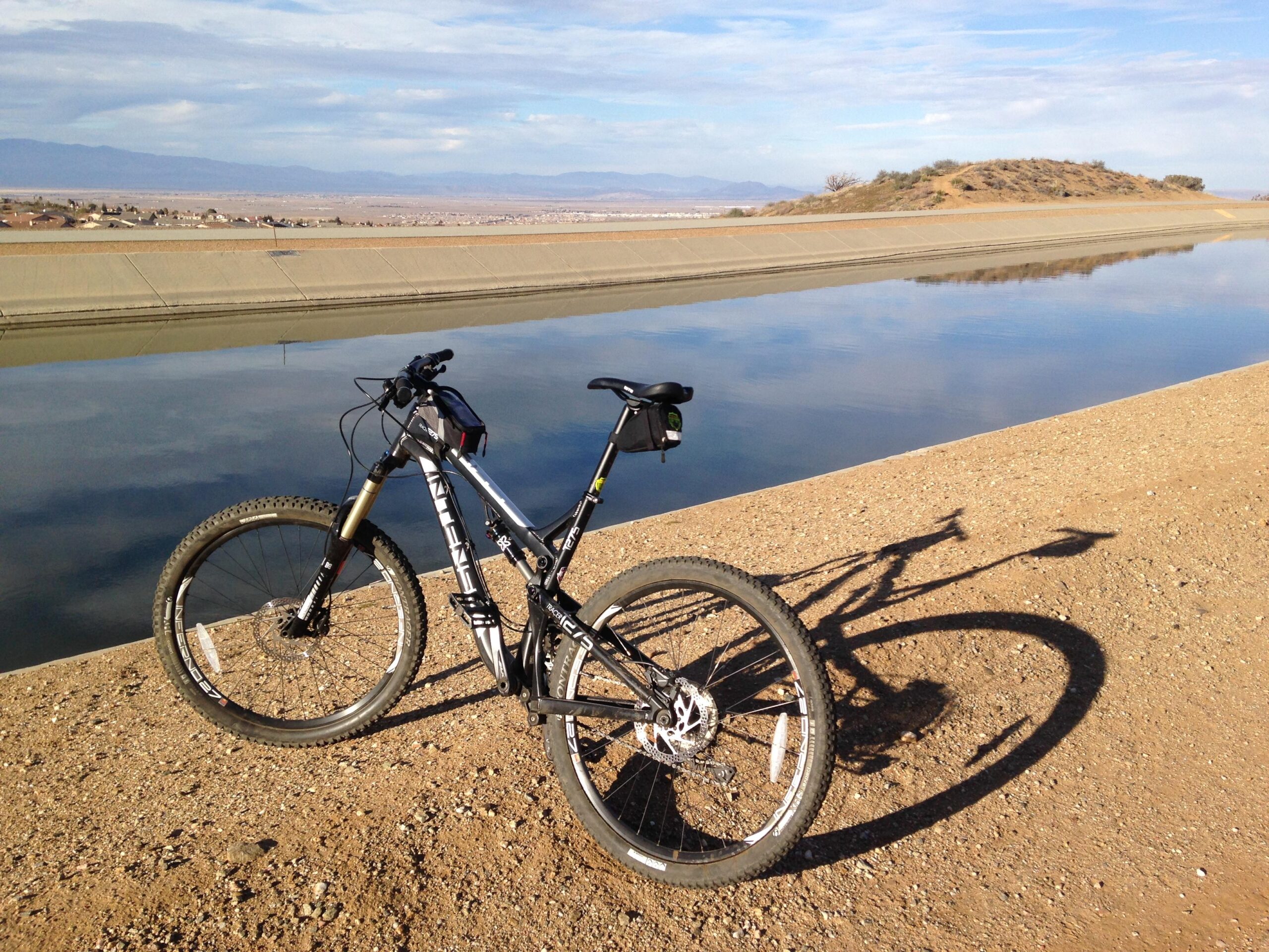 Intense tracer: A mountain bike parked on a sandy embankment beside a calm, reflective body of water, with distant mountains and a clear blue sky in the background.
