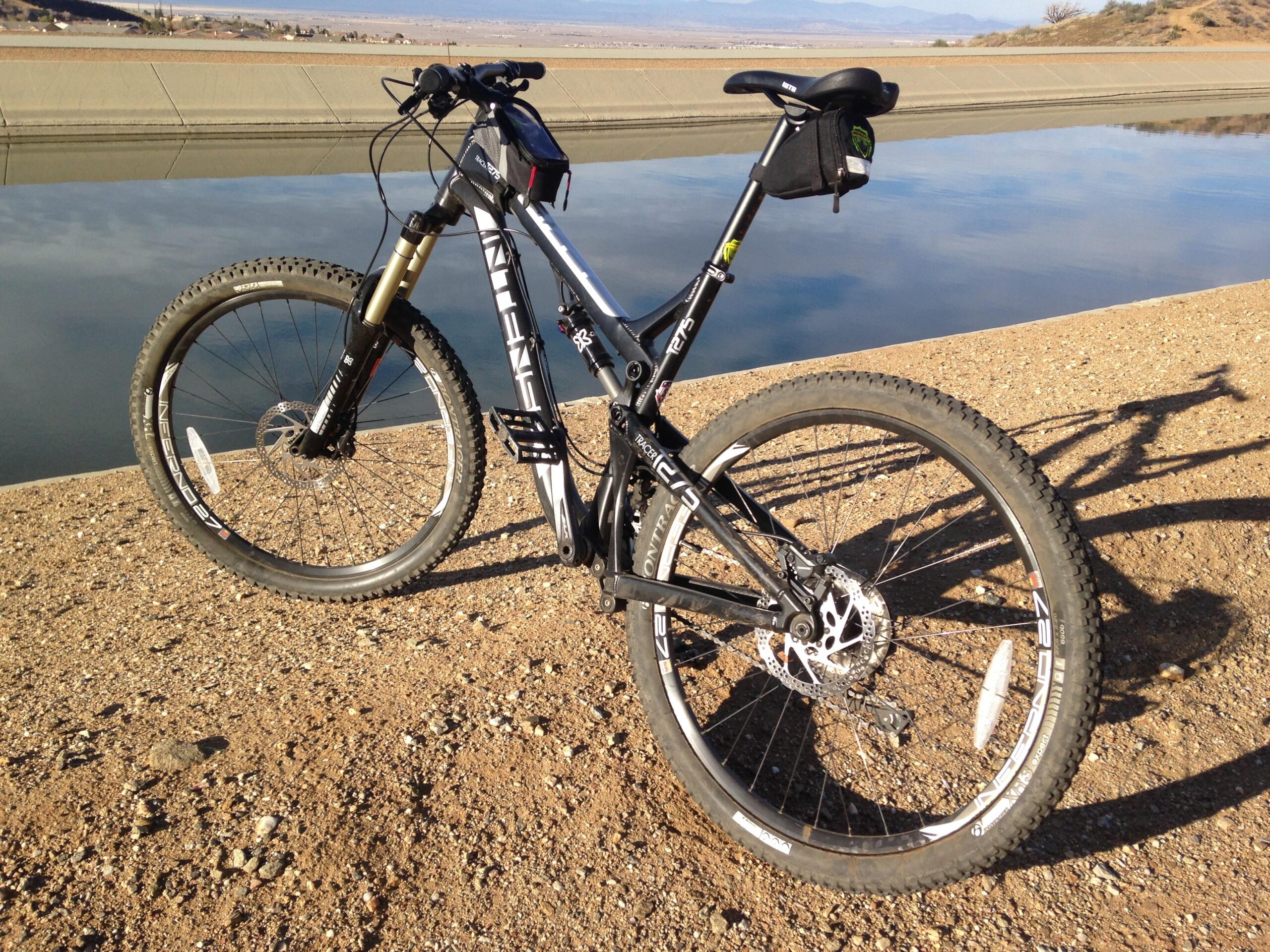 Intense tracer: A black mountain bike parked on a gravel path near a calm body of water, with a landscape of mountains in the background. The bike features prominent tires and a front suspension fork. Sunlight casts shadows on the ground, highlighting the bike's design and surroundings.