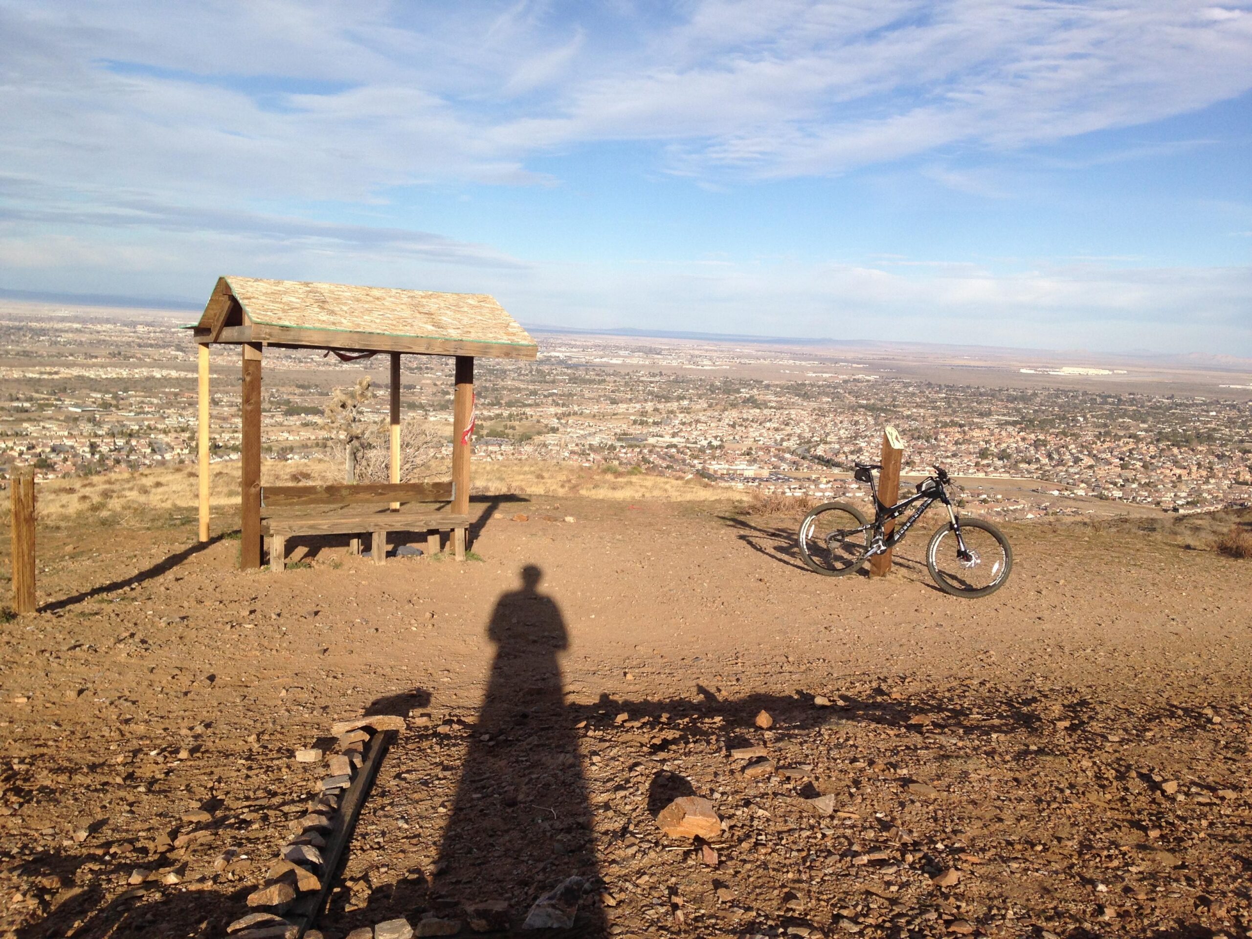 Intense tracer: A scenic view from a mountain overlook featuring a wooden shelter with a thatched roof, a mountain bike resting against a post, and a long shadow cast on the ground. The background showcases a sprawling landscape of fields and houses under a blue sky with some clouds.