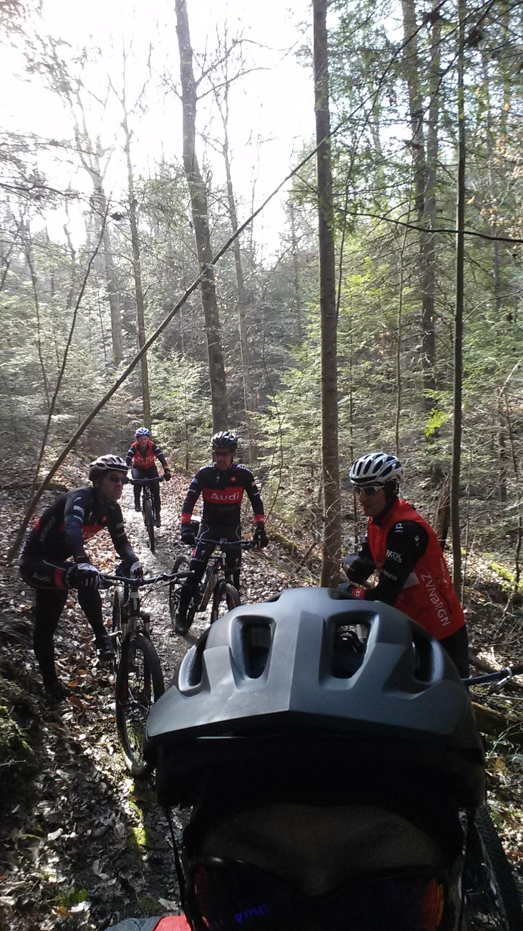 A group of four mountain bikers gather on a wooded trail, surrounded by trees and dappled sunlight filtering through the leaves. The foreground features a close-up of a cyclist wearing a black helmet and sunglasses, while the others are visible in the background, some wearing team jerseys. The scene conveys an active outdoor adventure in nature. Sheltowee Trace - Laurel Lake Trail mountain bike trail.