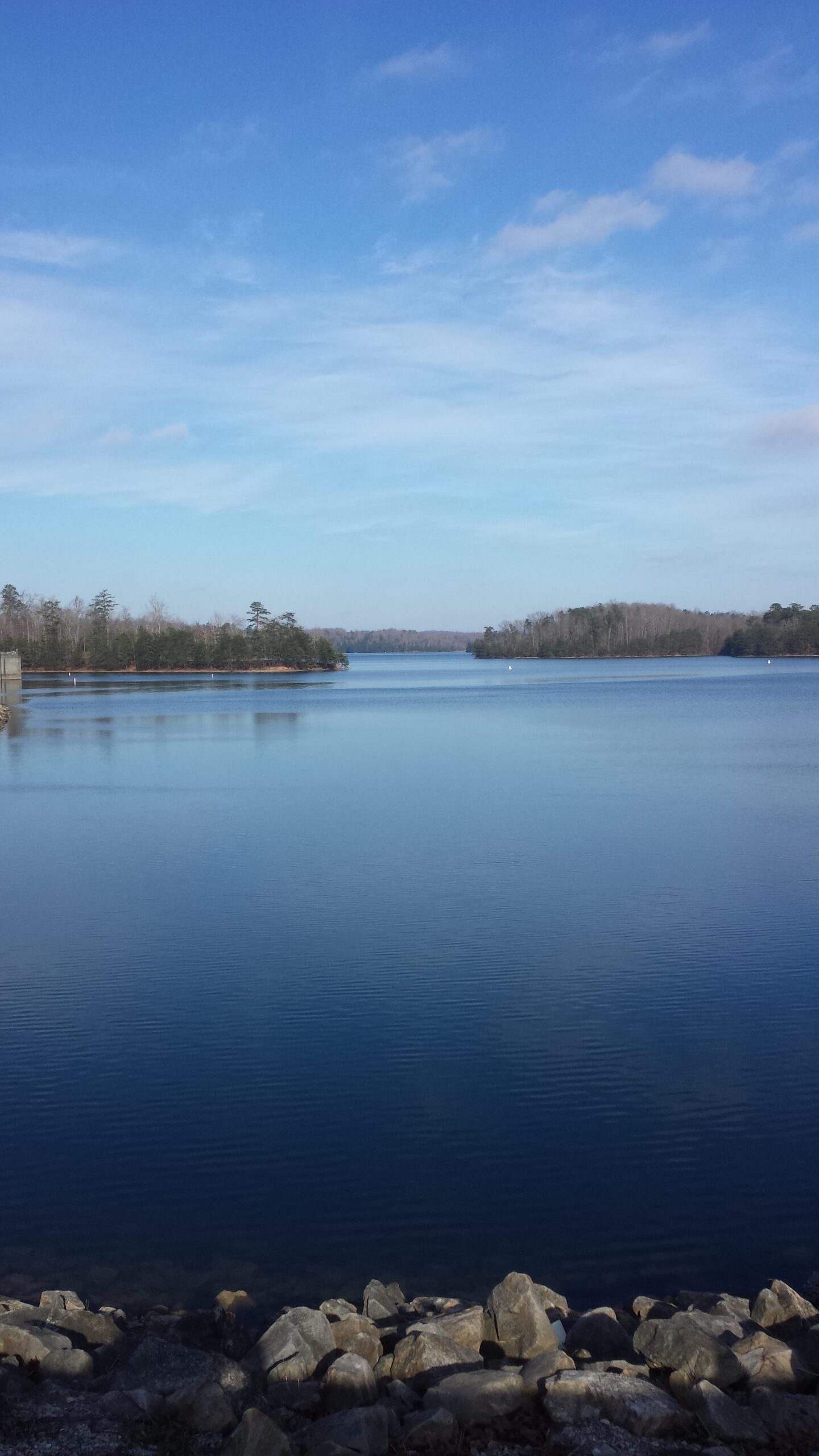 A tranquil view of a calm lake reflecting a clear blue sky. The foreground features smooth stones lining the water's edge, while trees can be seen along the shoreline, creating a peaceful, natural setting. Sheltowee Trace - Laurel Lake Trail mountain bike trail.