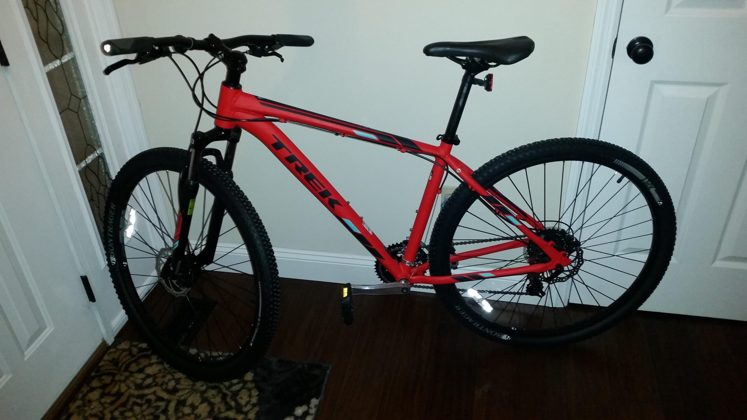 Trek Marlin 5: A red Trek mountain bike leaning against a wall, positioned in a well-lit indoor space. The bike features thick tires and a sturdy frame, with visible branding and reflective elements. A door and a patterned rug can be seen in the background.