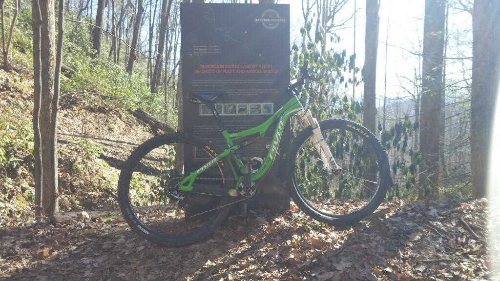 Pivot Mach 429 Carbon: A green mountain bike parked beside an information sign in a wooded area. The background features tall trees and a leaf-covered ground, suggesting a natural trail environment. The sign includes text and graphics related to local plant and animal diversity.