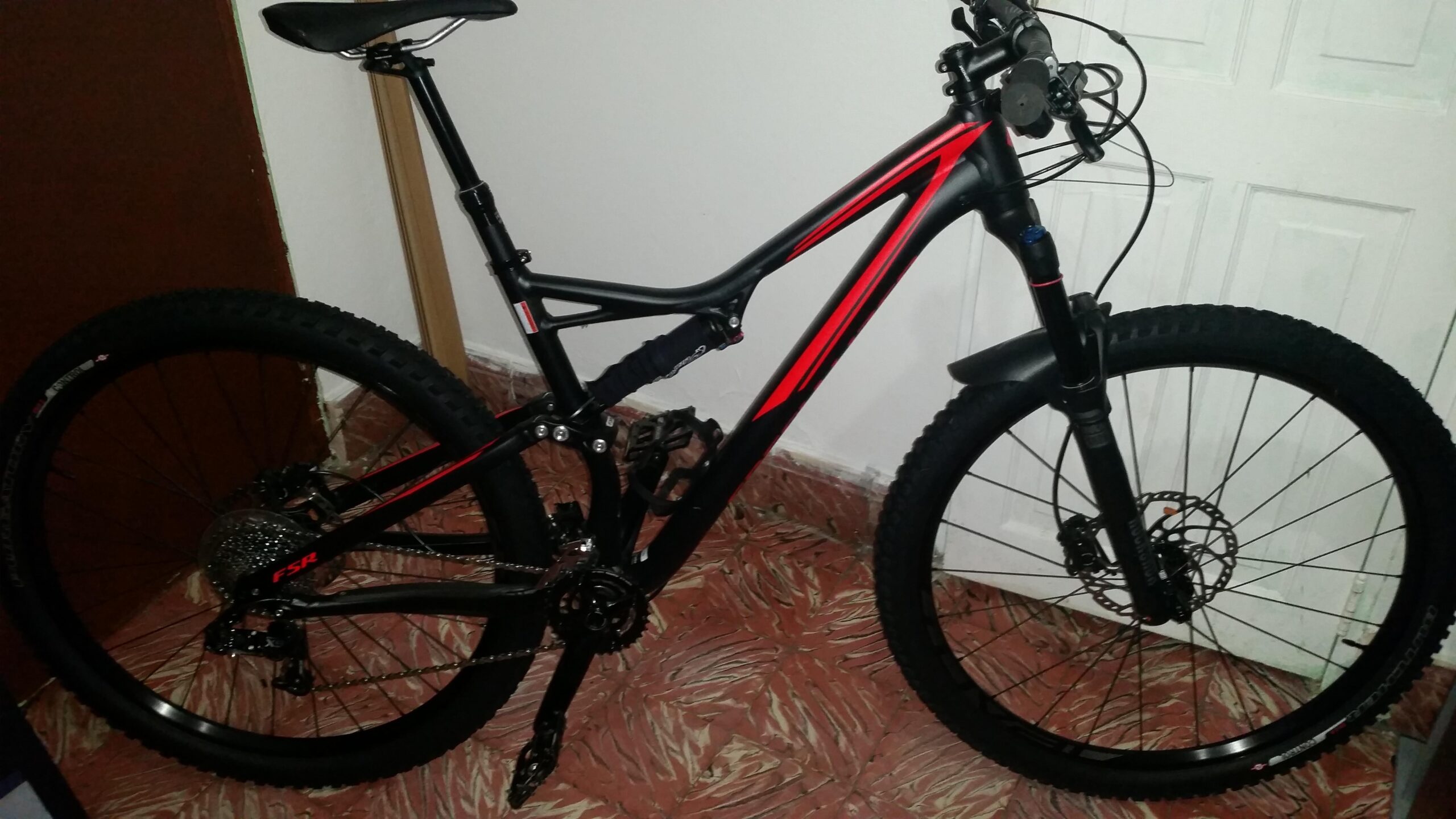 Specialized Stumpjumper FSR: A black and red mountain bike is leaning against a wall with a closed door in the background. The bike features thick tires, a front suspension fork, and a dropper seat post. The floor has a patterned design, adding a warm tone to the setting.