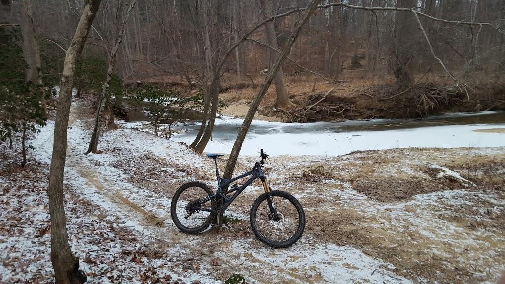 A mountain bike parked on a snowy trail next to a frozen creek, surrounded by bare trees in a wooded area during winter. The scene captures the tranquility of nature in a cold environment. Cabin John Trail mountain bike trail.