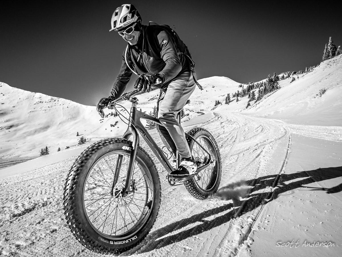 A person riding a fat bike on a snowy trail, wearing a helmet and sunglasses, with snow-covered mountains in the background. The image is in black and white, emphasizing the contrast between the rider and the snow. Cottonwood Pass Road mountain bike trail.