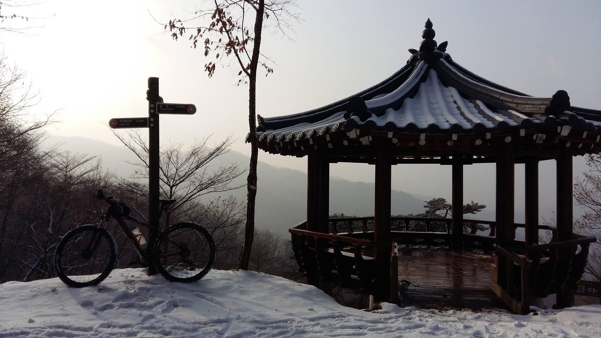 A scenic winter landscape featuring a wooden gazebo with a traditional roof, surrounded by a blanket of snow. A mountain bike is parked nearby on the snowy ground. In the background, misty mountains are visible, and a signpost with directional arrows is positioned next to the gazebo, indicating various distances to nearby locations. The overall ambiance is serene, with soft lighting from the sun peeking through the fog. Wangbangsan mountain bike trail.