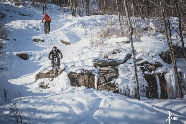 Two mountain bikers navigating a snowy landscape, with one cyclist descending a rocky section while the other rides slightly behind on a snow-covered trail. Icicles hang from the rocks in the background, and trees with bare branches line the scene, indicating a winter setting.