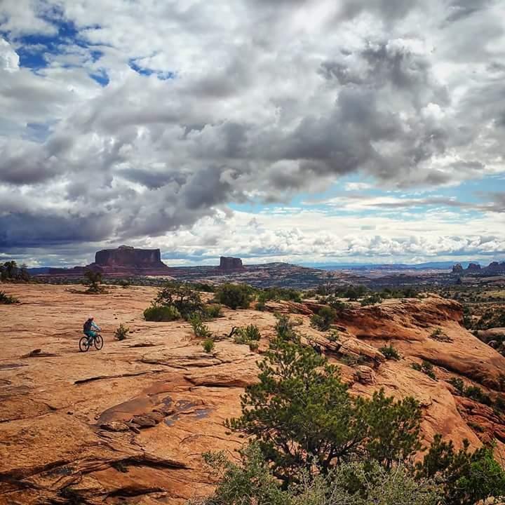 A person riding a mountain bike on rocky terrain, with expansive views of distant mesas and a cloudy sky in the background. Vegetation is present among the rocks, showcasing a rugged, natural landscape. Navajo Rocks mountain bike trail.