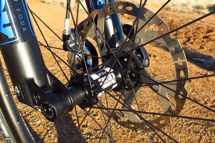 Close-up view of a mountain bike front wheel, focusing on the disc brake system, which includes a rotatable metallic disc and a black hub, set against a sandy background. The bike frame features bold blue graphics.